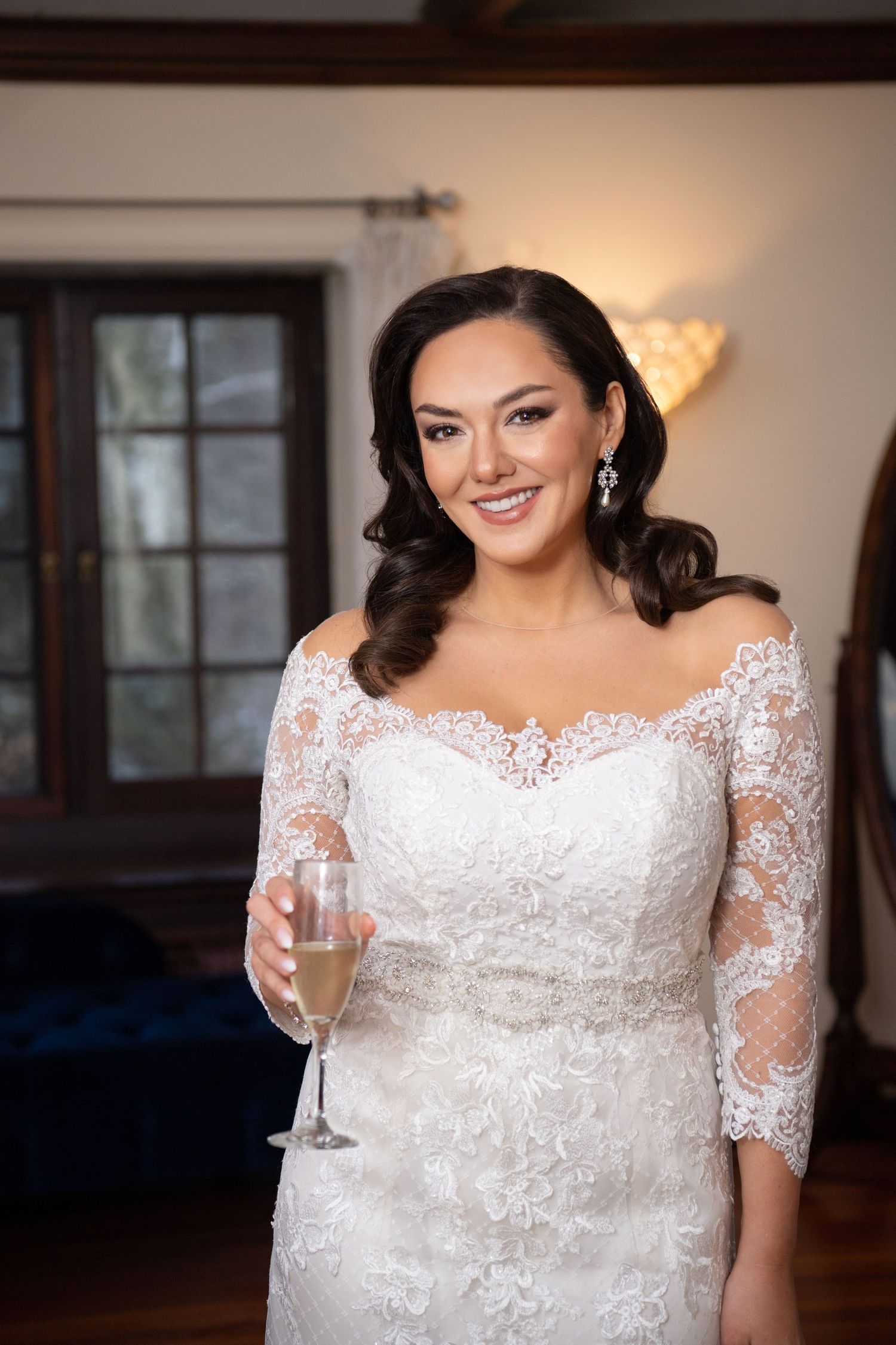 A bride in a white lace wedding dress with three-quarter length sleeves holds a champagne glass while smiling.