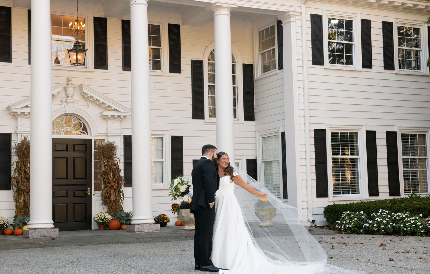 Elegant white colonial style mansion with pillars and landscaping serves as backdrop for wedding couple.