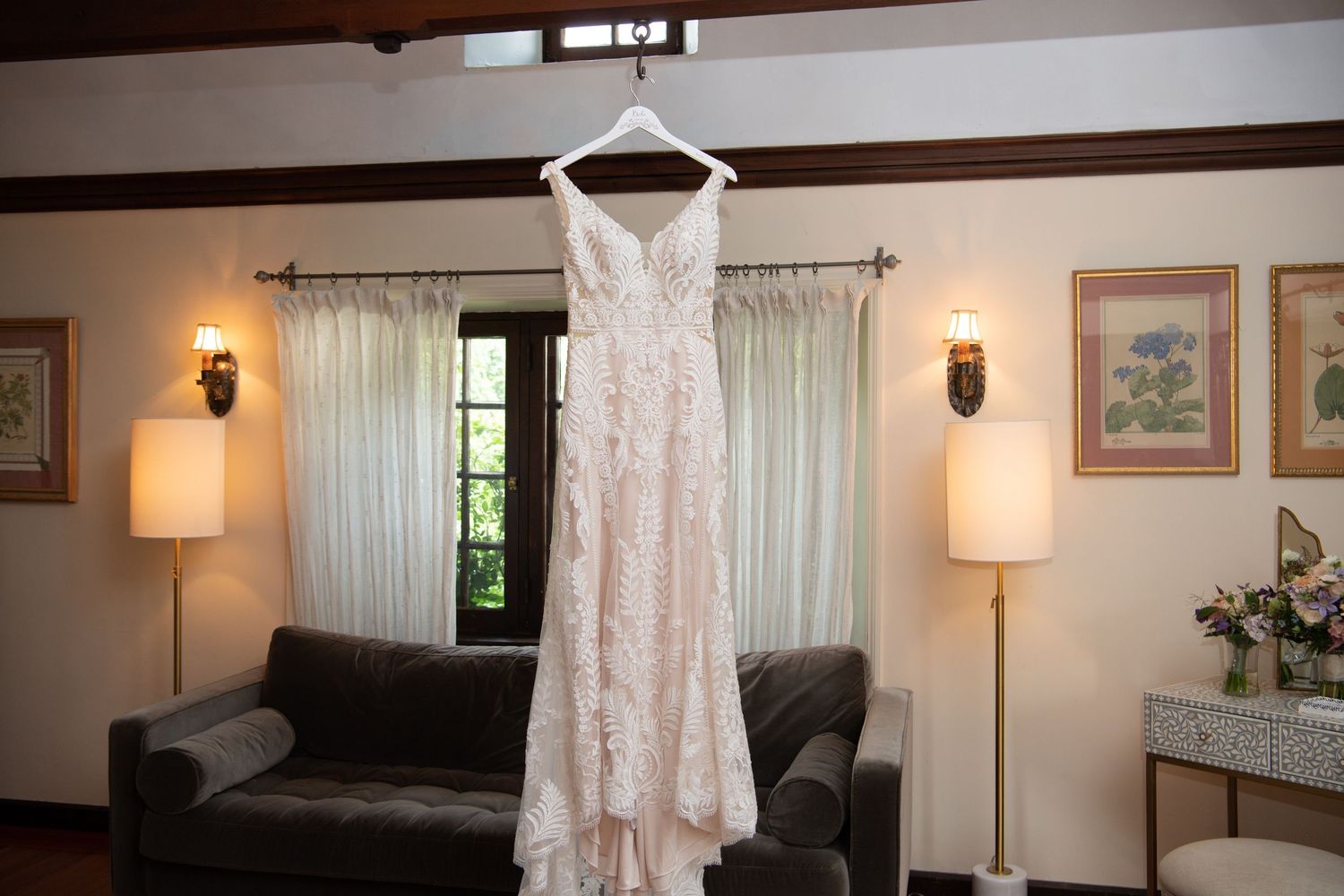 A delicate white lace wedding dress hangs elegantly on a hanger in a cozy room with table lamps and curtained windows.