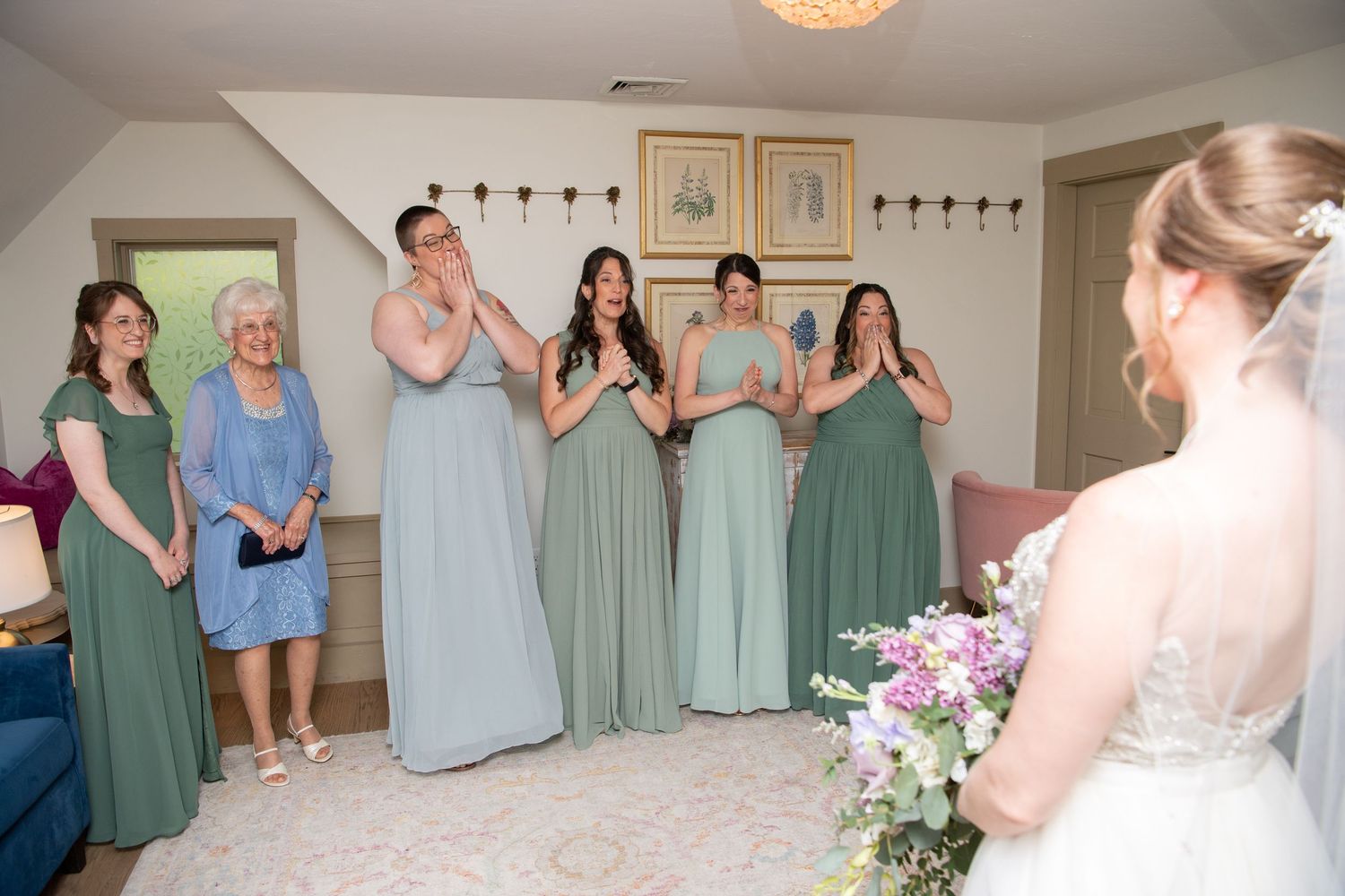 A wedding party getting ready in a room with sage green and light blue dresses and floral bouquets.