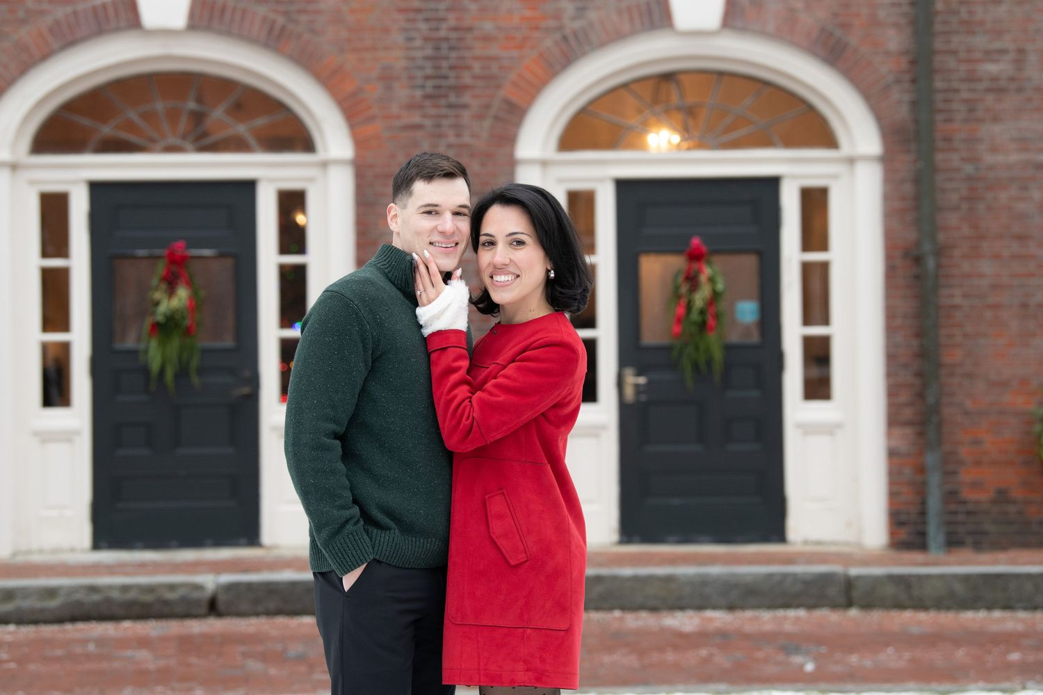 A couple in formal winter attire poses for photos outside a historic brick building decorated with holiday wreaths.