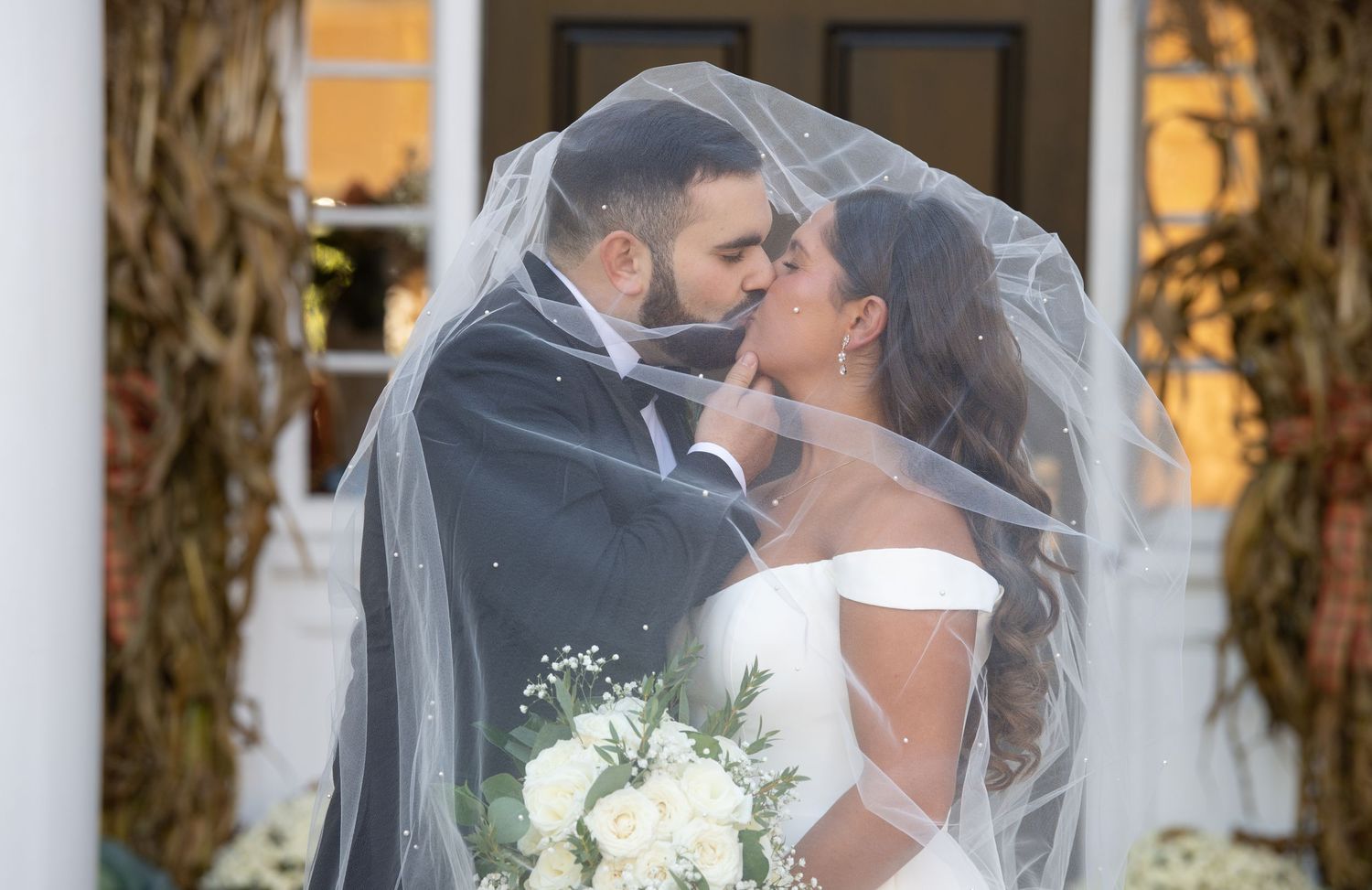 A romantic wedding moment captured under a veil as a couple shares a kiss while holding a white floral bouquet.