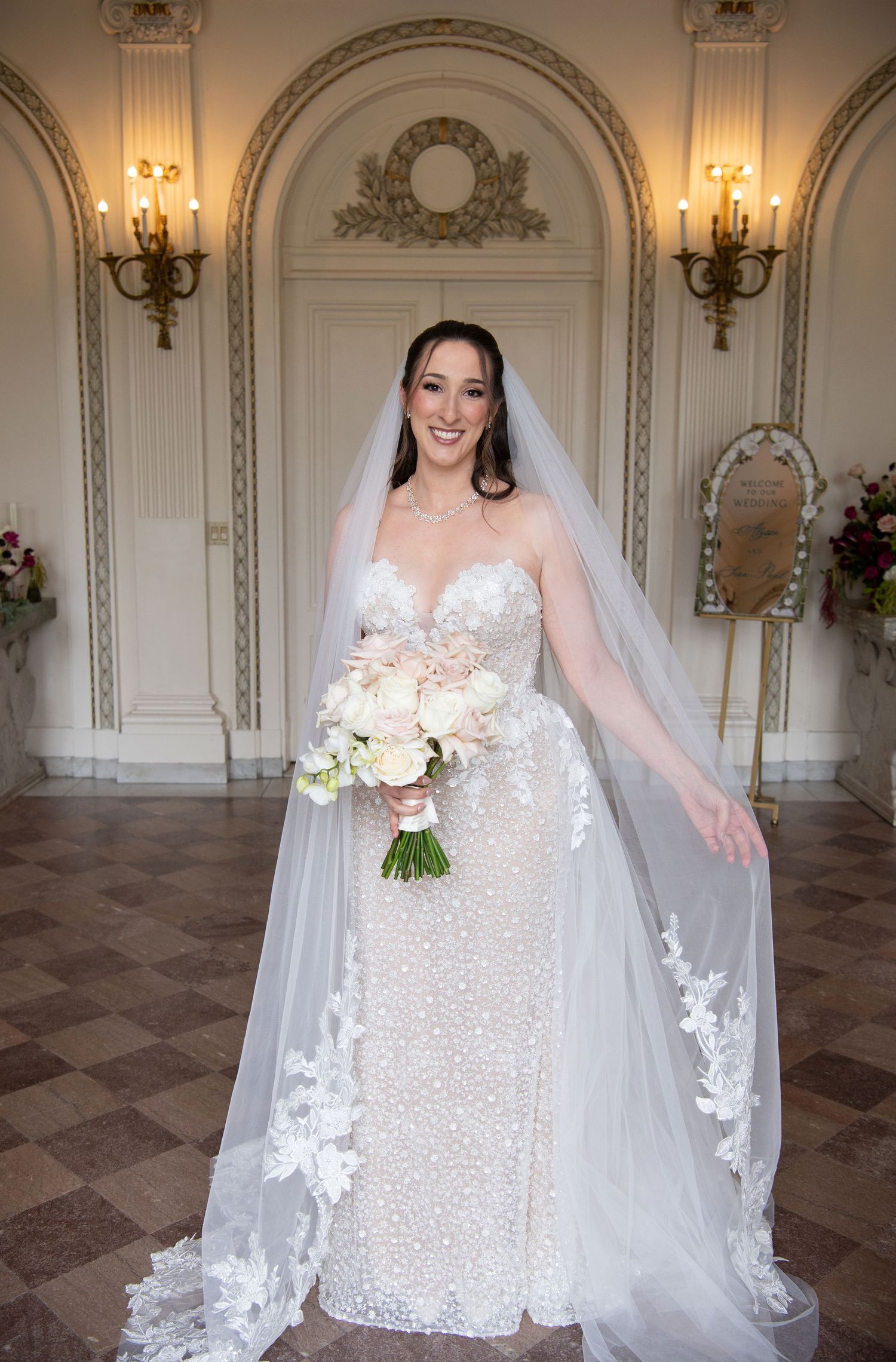 Smiling bride in pearl-embellished gown with lace veil holds bouquet in elegant gold-accented ballroom.