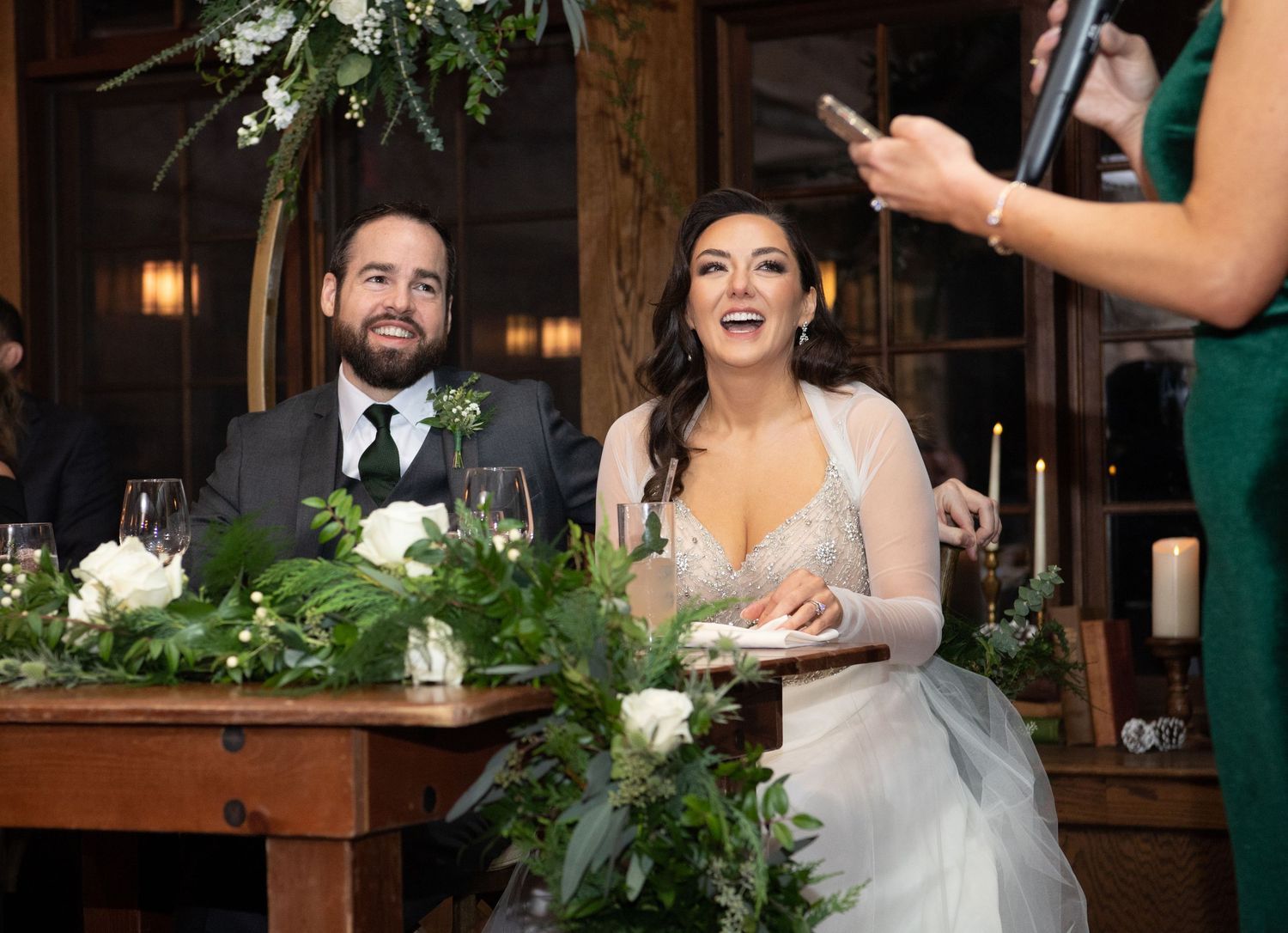 A happy couple shares a joyful moment during their wedding reception in a rustic venue decorated with greenery.