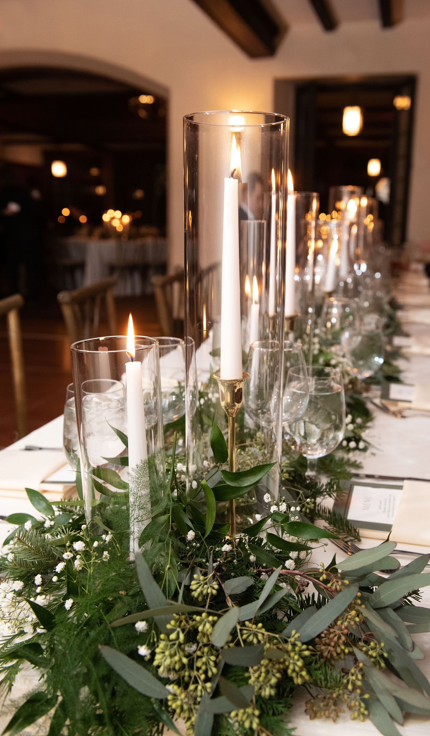 Elegant table setting with tall glass candle holders and lush greenery garland centerpiece for a wedding reception.