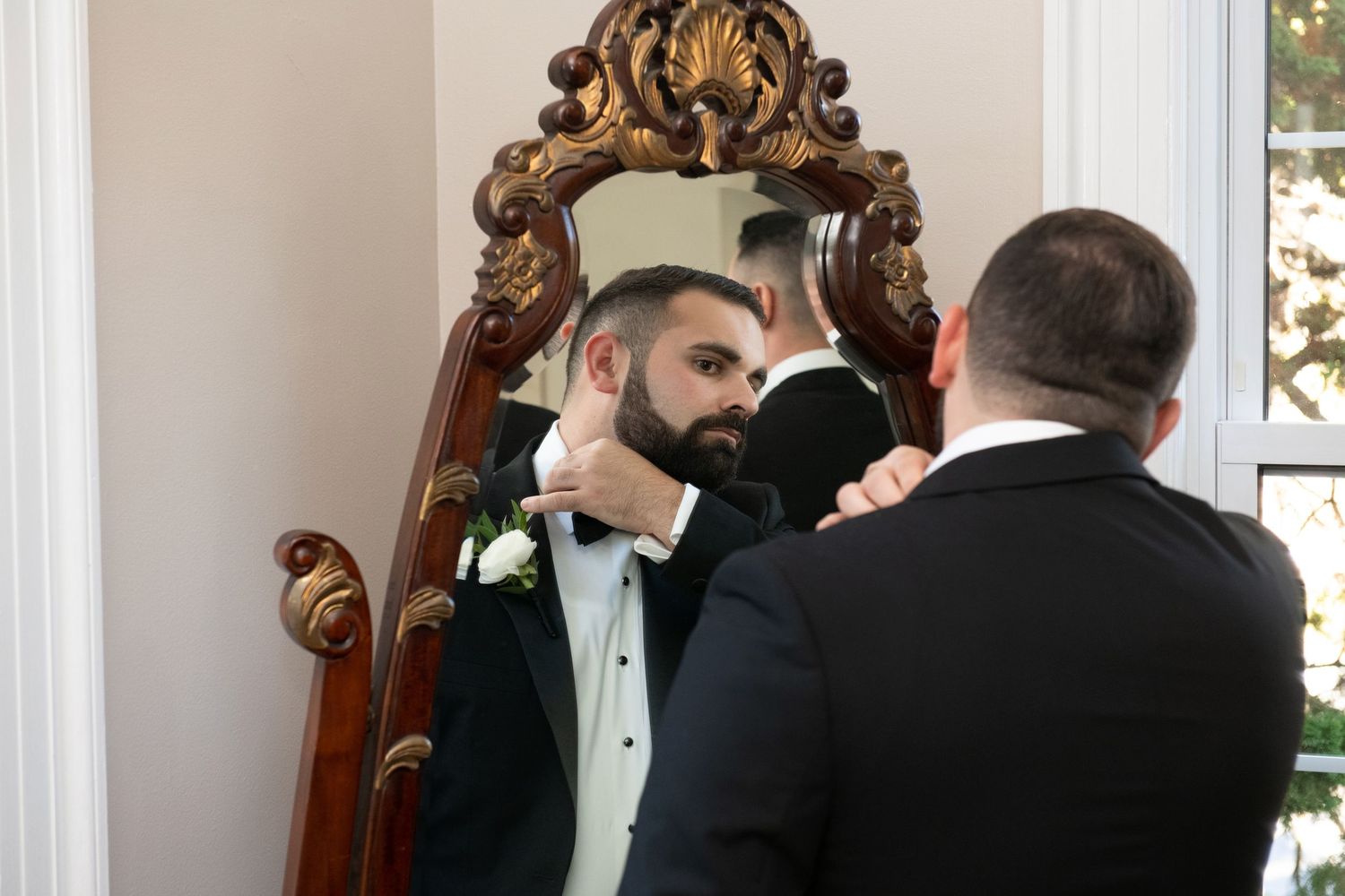 A group of formally dressed people gather around an ornate mirror in an elegant white-walled room.