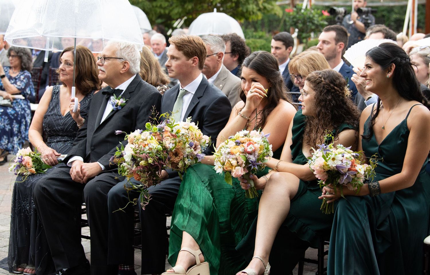 Wedding guests seated outdoors in formal attire with bridesmaids wearing emerald green dresses.