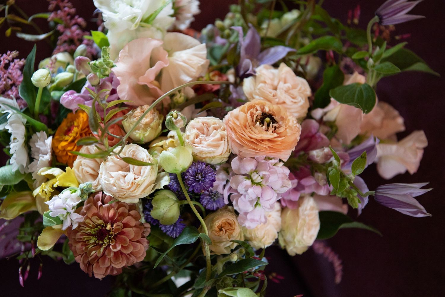 Lush floral arrangement featuring peach ranunculus, purple blooms, and mixed greenery in moody lighting.