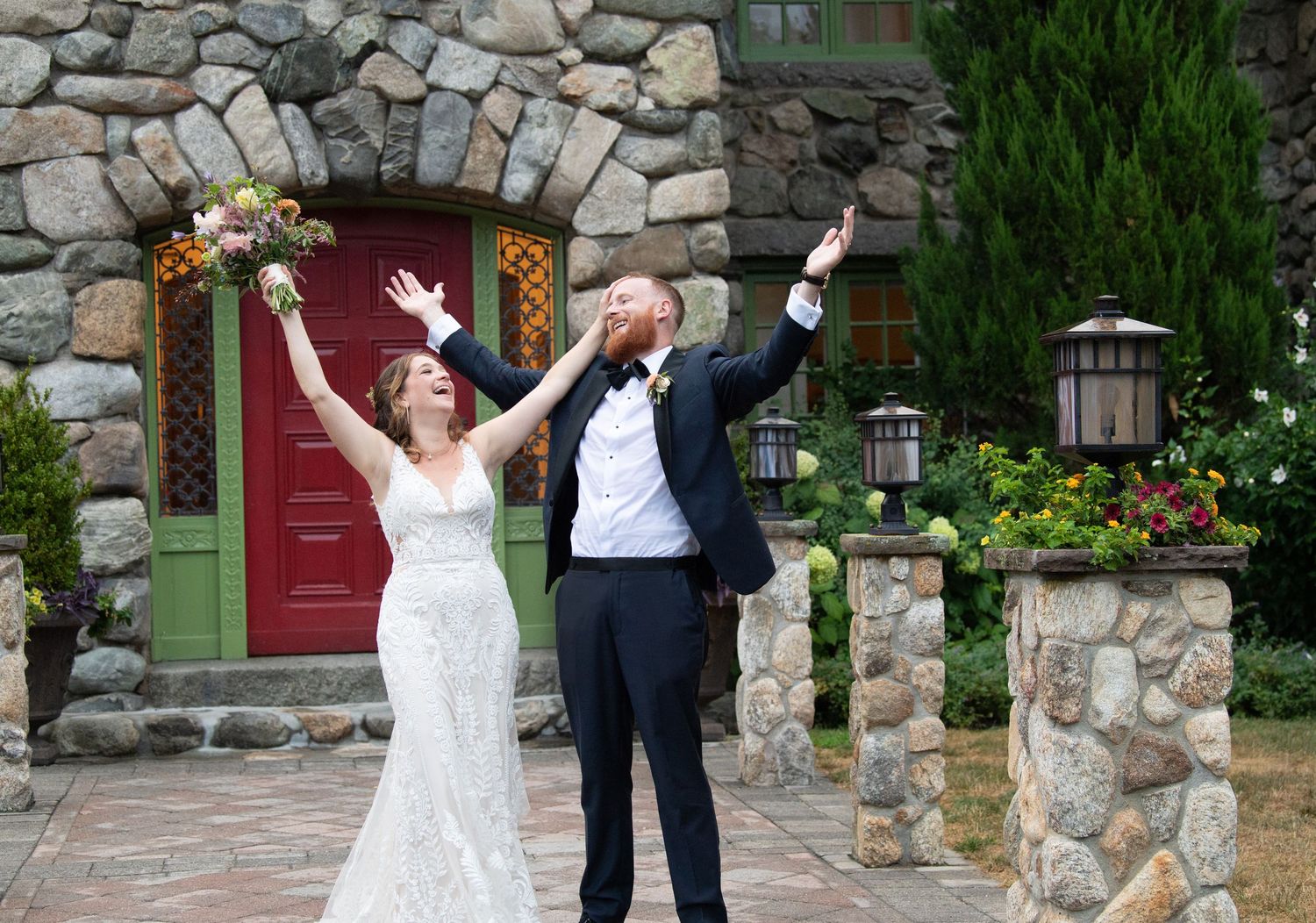 Joyful couple celebrates with champagne in front of rustic stone archway with red door and garden lanterns.