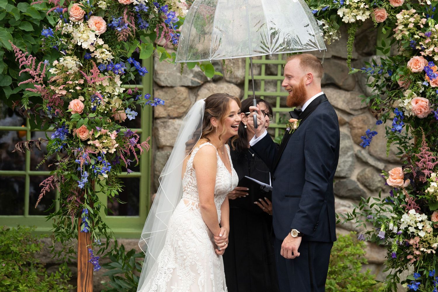 A wedding couple embraces under a romantic floral arch with pink roses and greenery in a garden setting.