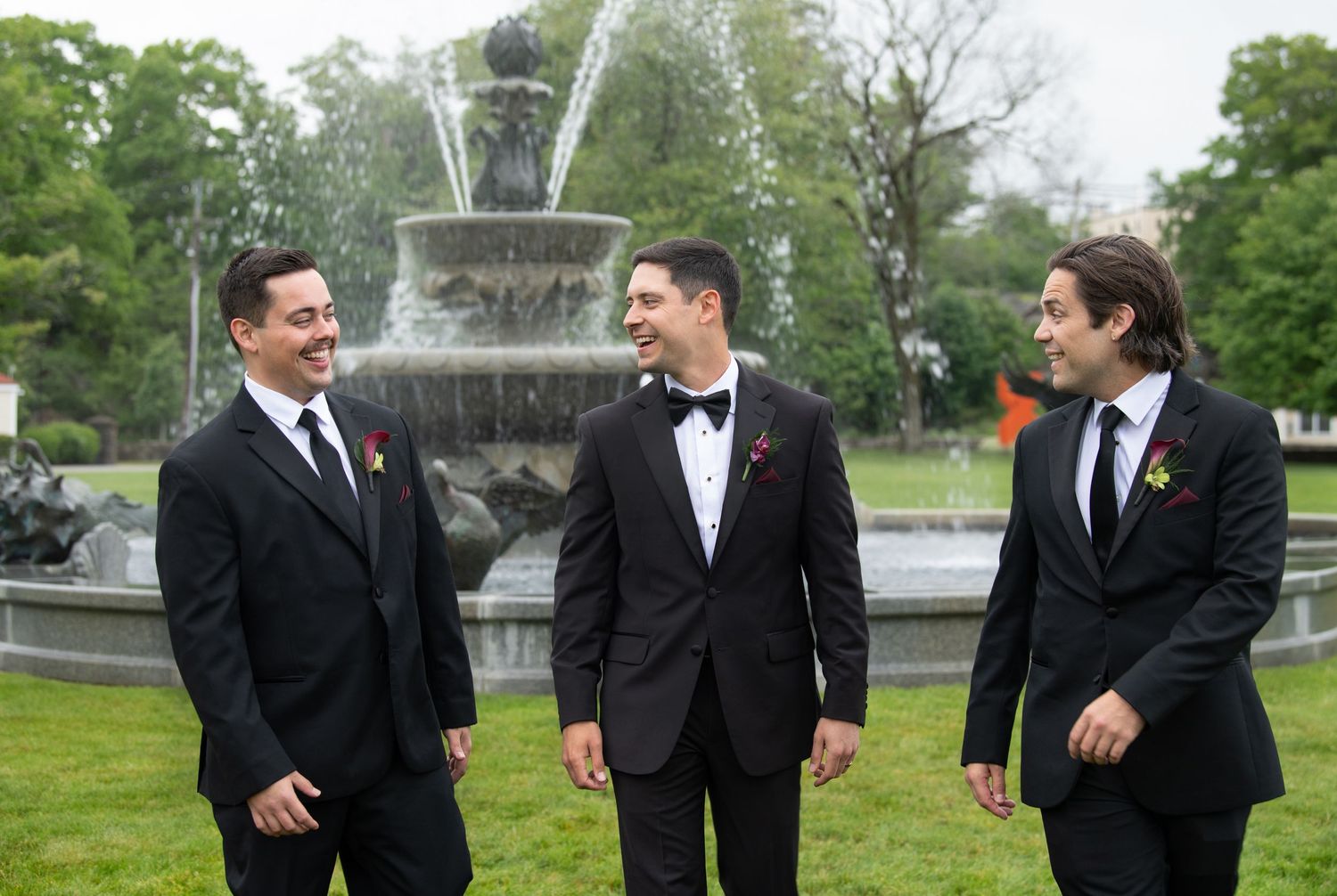 Three groomsmen in black tuxedos laughing together outdoors near an ornate stone fountain in a park.