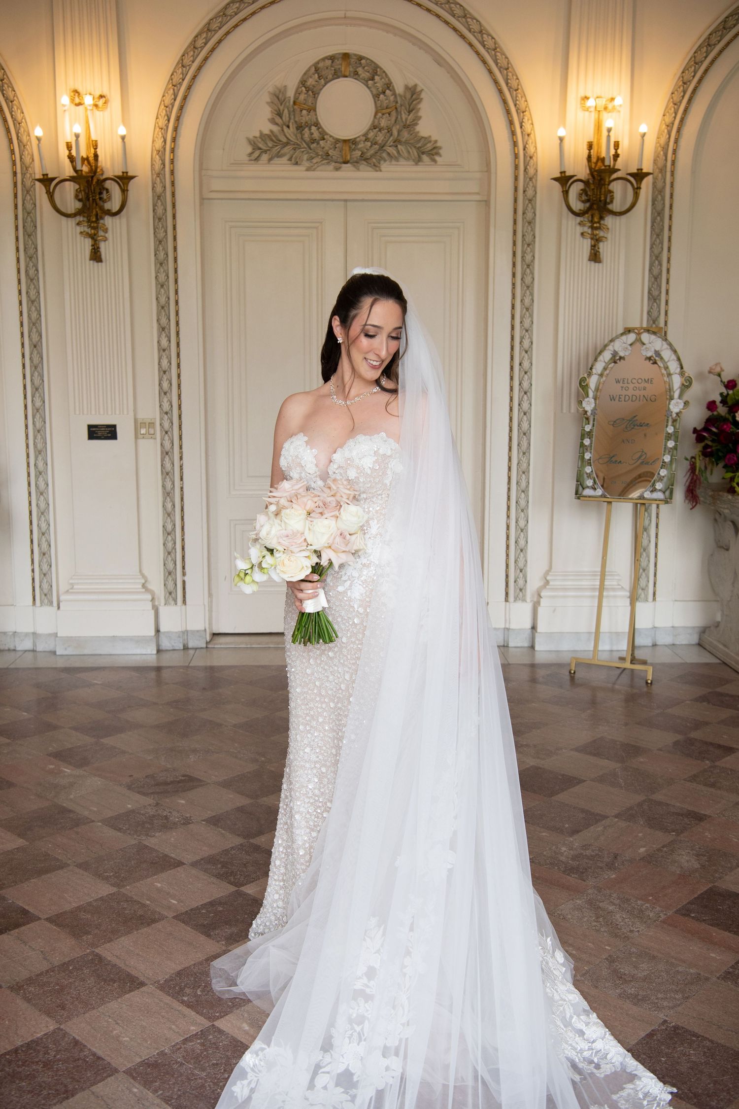 Bride in beaded strapless gown with veil holds white bouquet in ornate ballroom.
