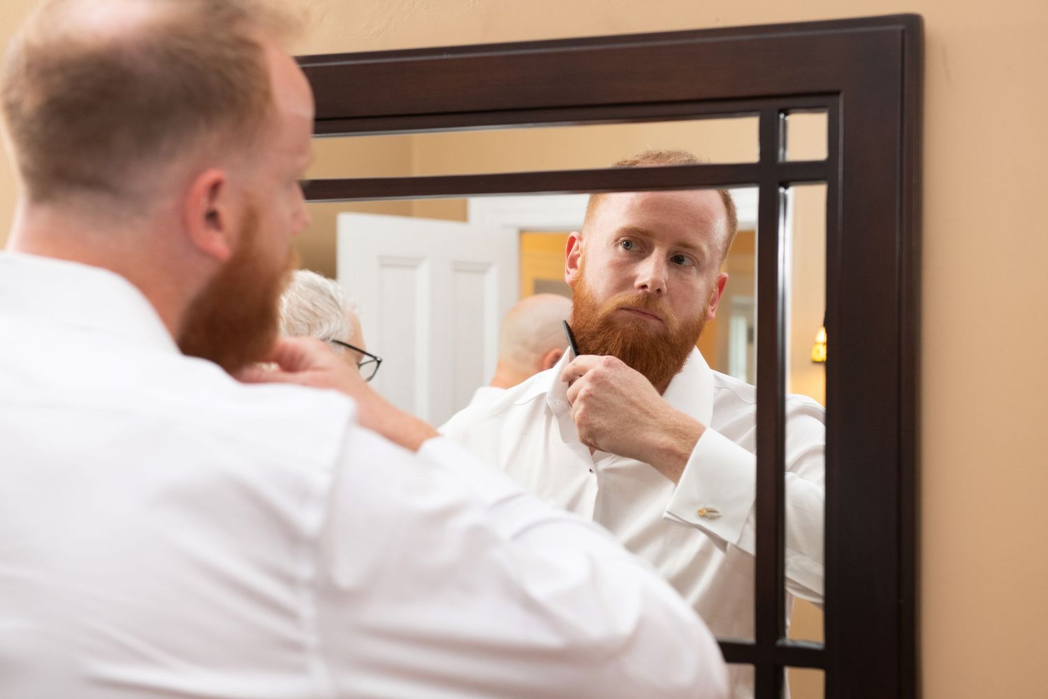 A person in a white shirt grooms their red beard while looking in a mirror.
