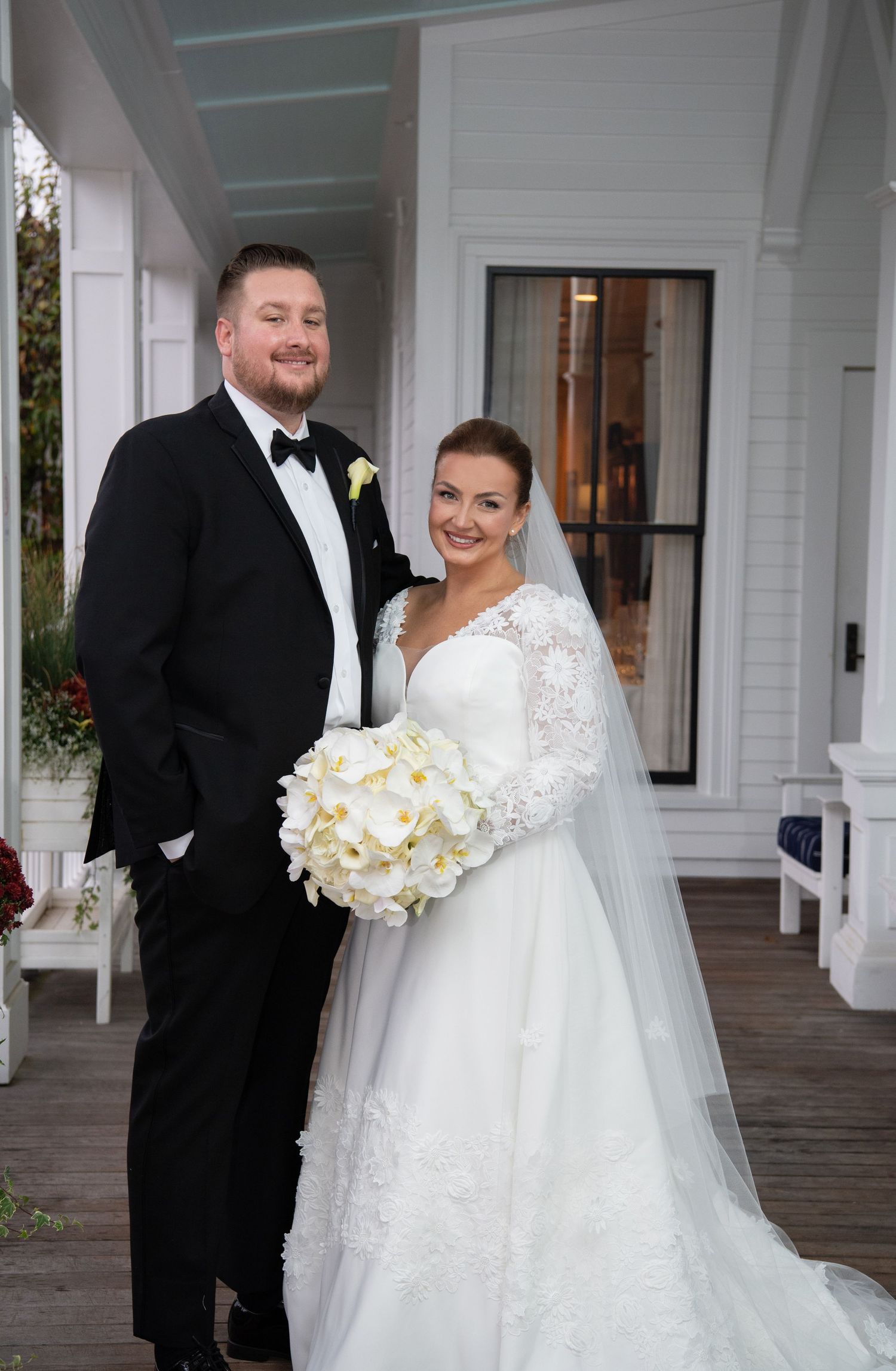 A wedding couple poses on a white columned porch with the bride holding a bouquet of white flowers.