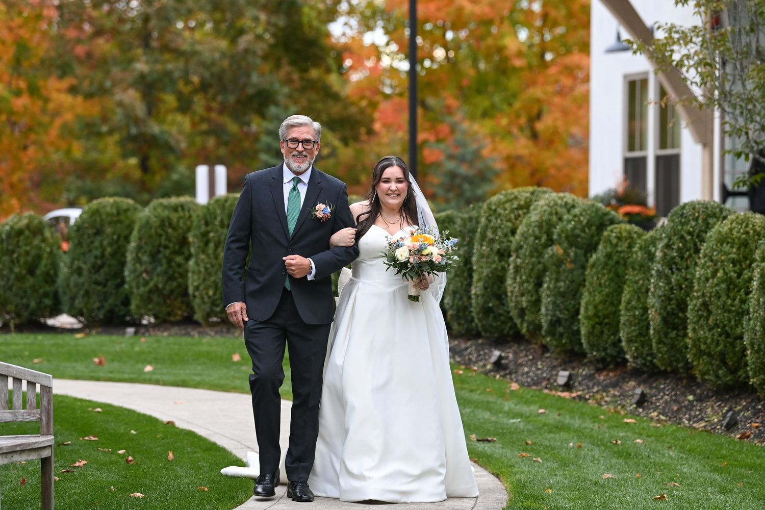 Wedding party walks along a manicured garden path with green hedges during autumn.