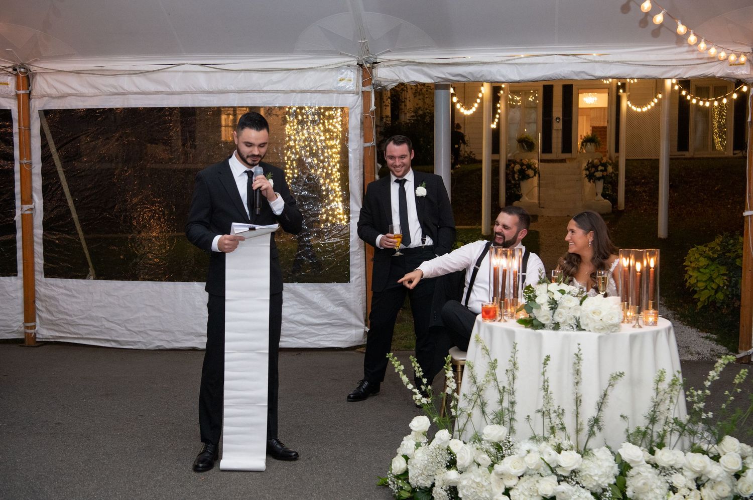 Wedding reception speech under a white tent with string lights and elegant floral decorations at an evening celebration.