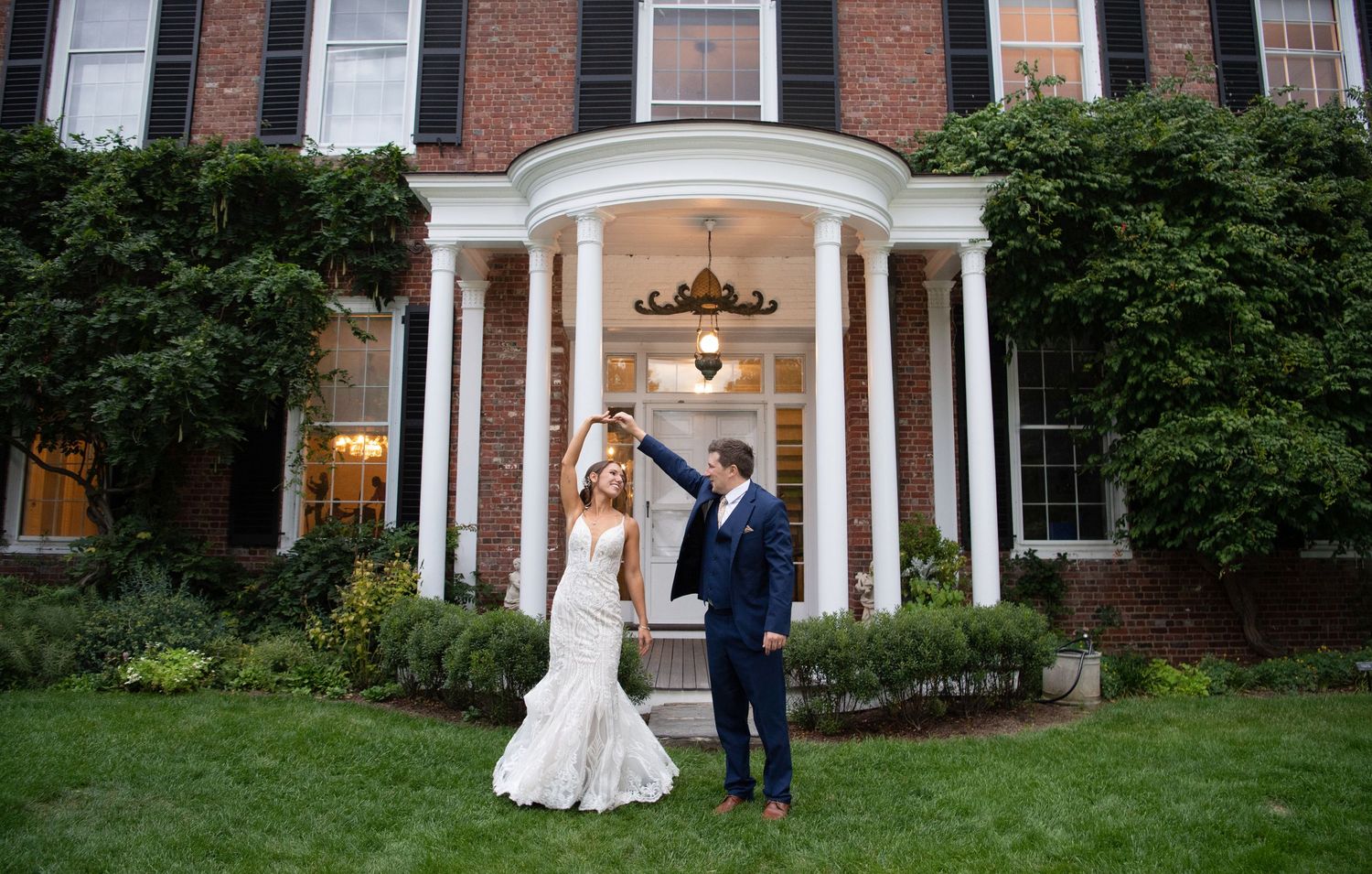 Newlyweds pose under elegant white columned portico of brick mansion.