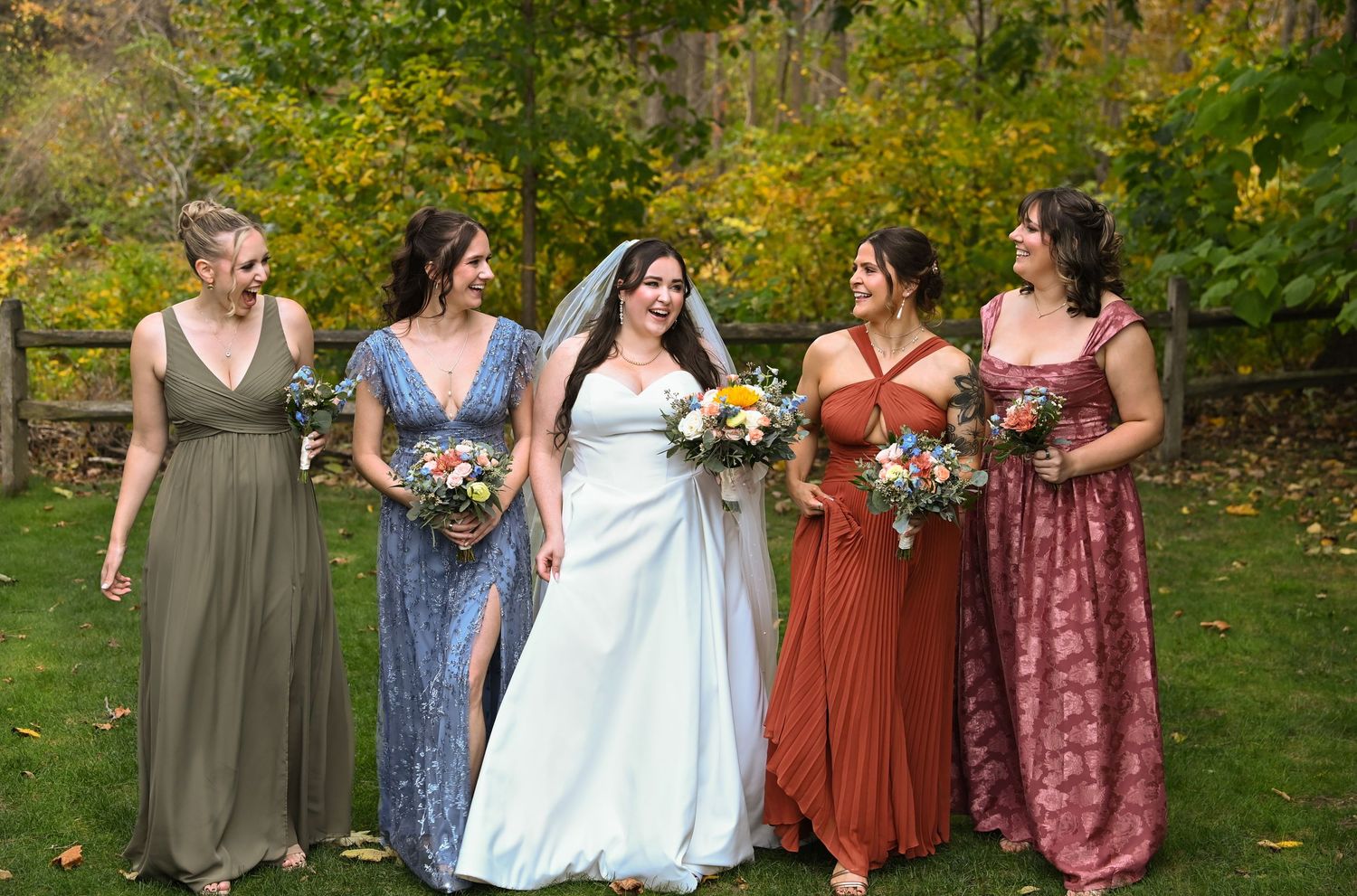 A wedding party in autumn poses outdoors wearing coordinated dresses in shades of burgundy, blue and olive green.