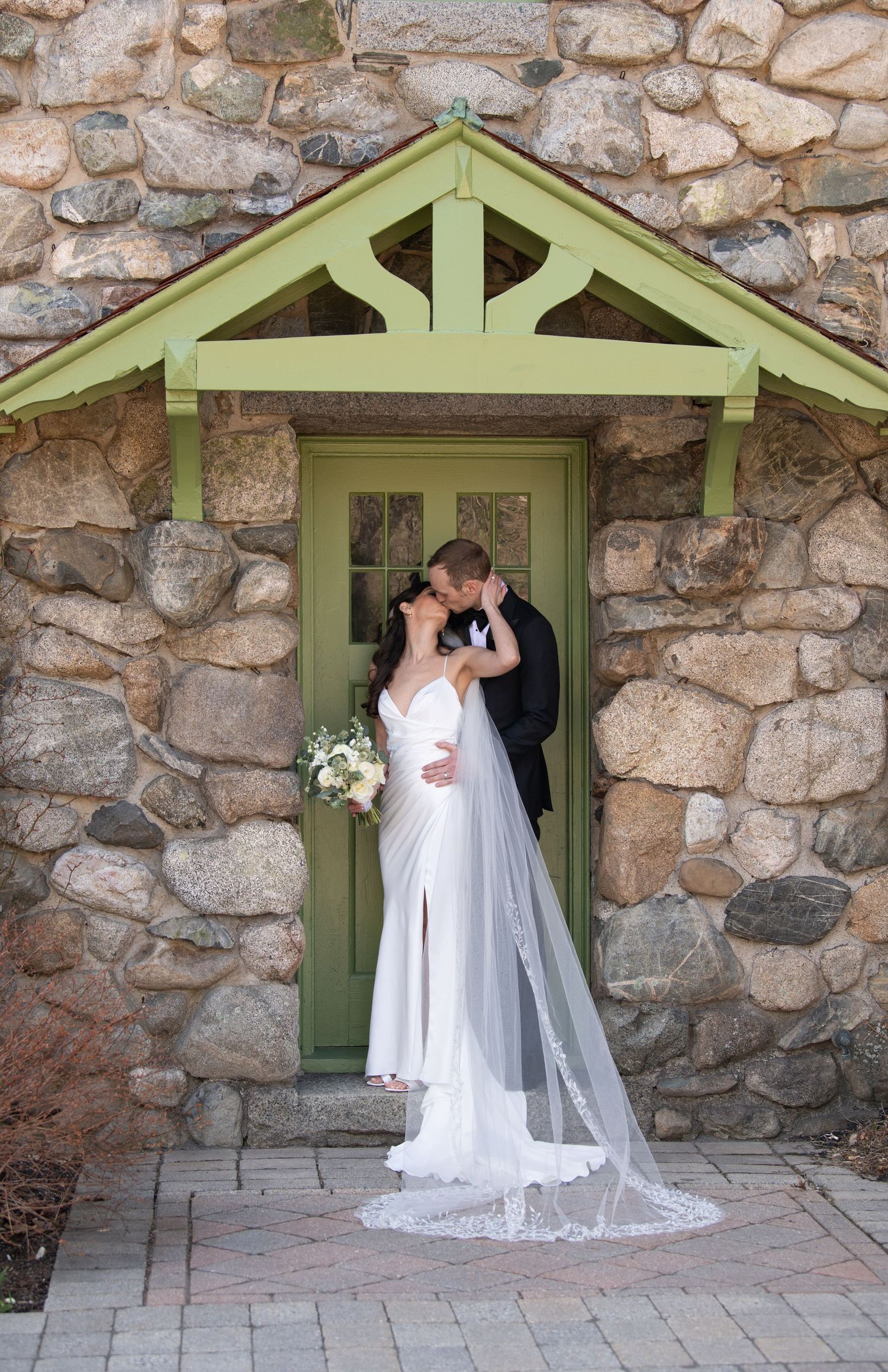 Bride and groom kissing in front of a green door set in a rustic stone wall, bride wearing a flowing white gown with veil.