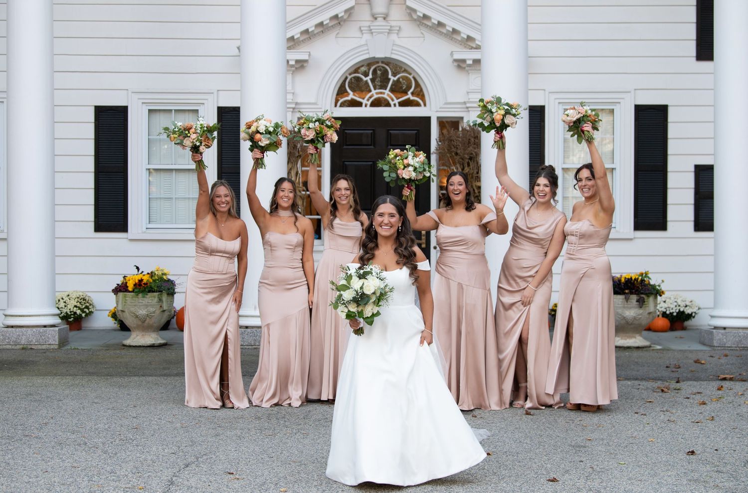 Wedding party in blush pink bridesmaid dresses poses together in front of white colonial style building.