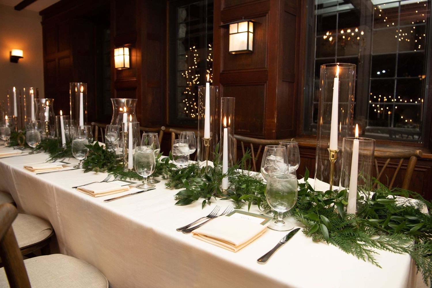An elegant table setting with tall candles and evergreen garland runners on a white tablecloth.