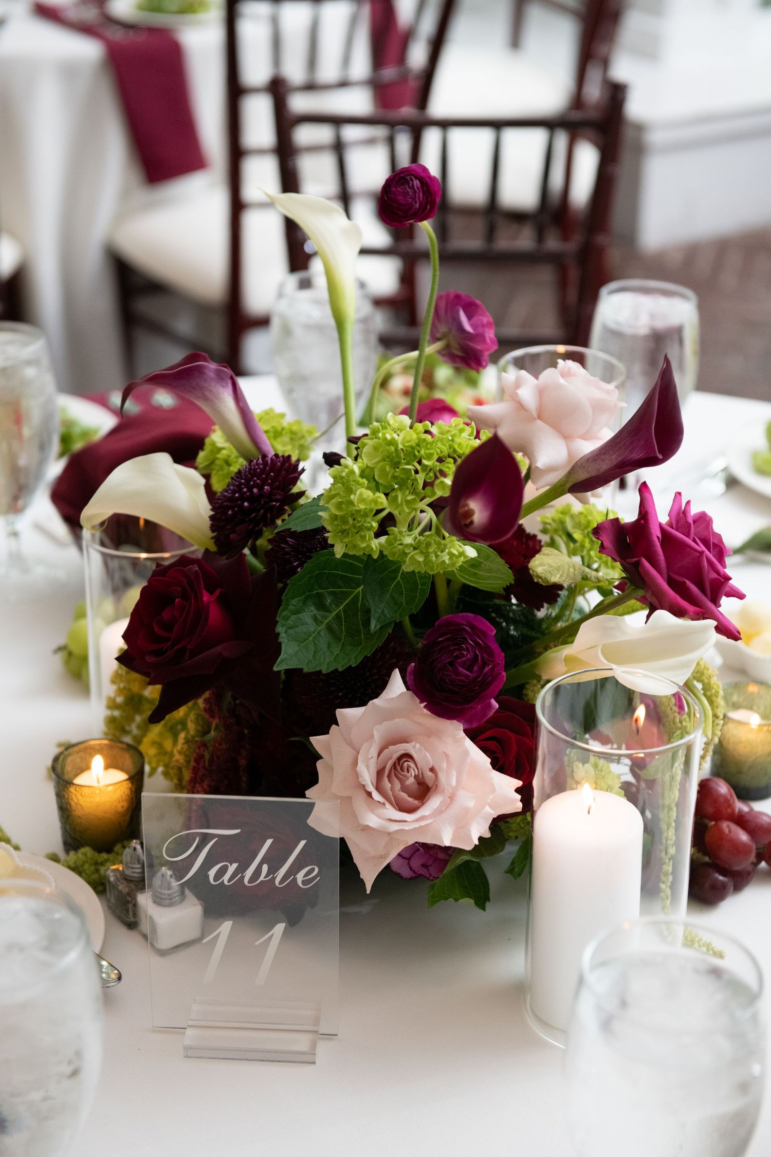 Elegant wedding table centerpiece with burgundy roses, calla lilies, and green hydrangeas beside a Table 11 sign and candles.