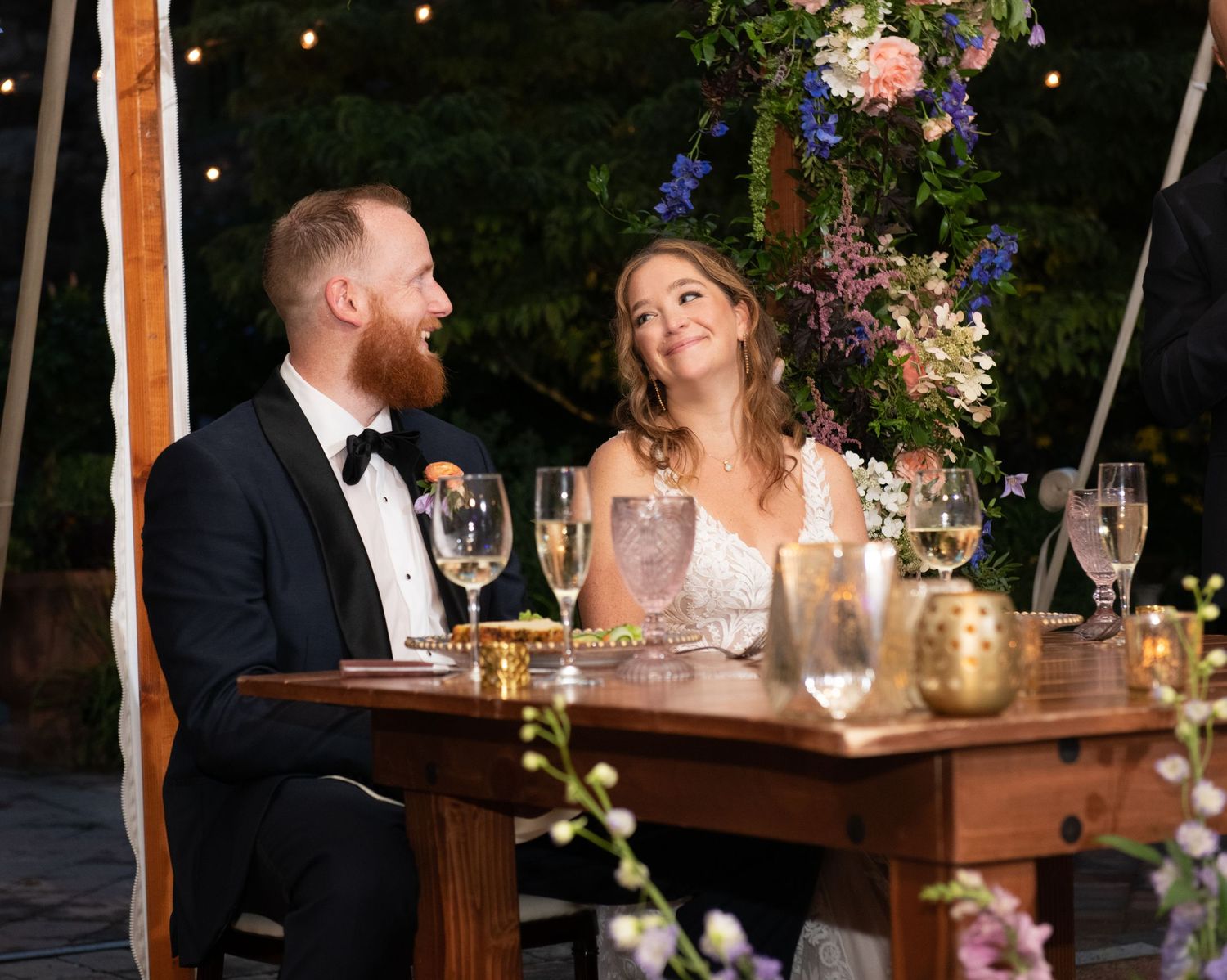 Romantic couple sitting at wooden table under string lights with floral arrangements at outdoor wedding reception.