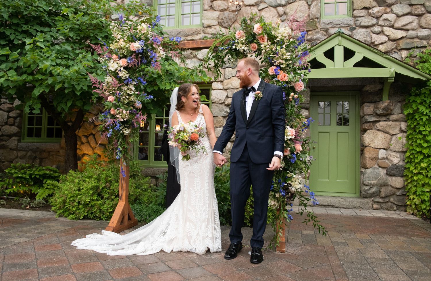 A wedding ceremony under a floral arch with stone walls and a green door in the background.