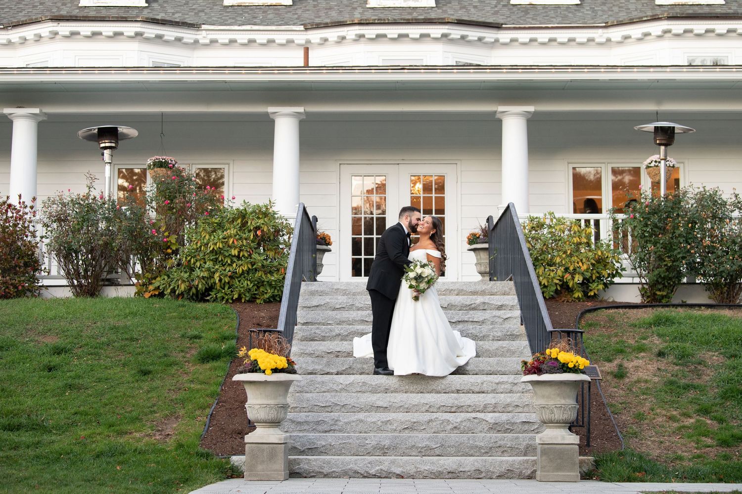 A couple stands on grand white steps leading to an elegant mansion with columns and French doors decorated for a wedding.