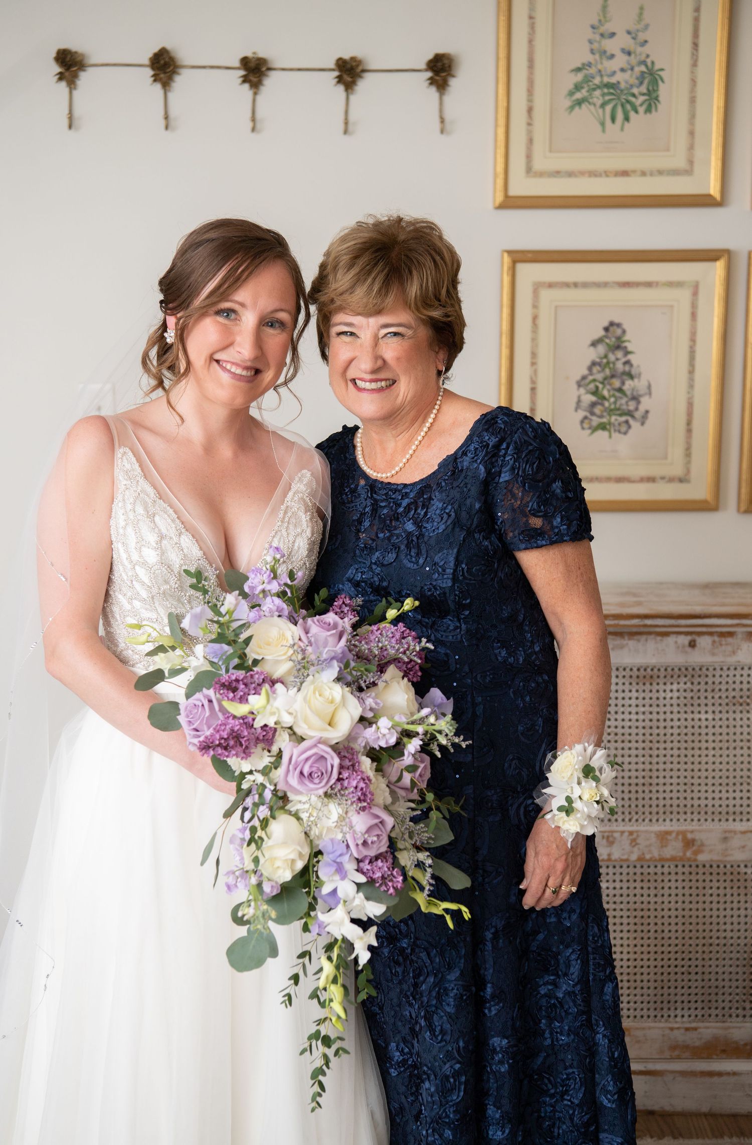 A bride in white gown poses with someone in navy blue lace dress holding a purple and white bouquet.