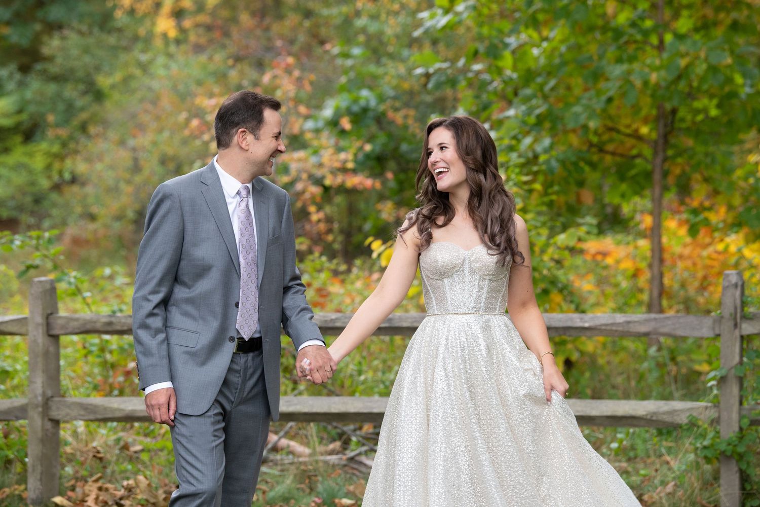 Bride and groom holding hands smiling at each other in front of a wooden fence with autumn foliage backdrop.
