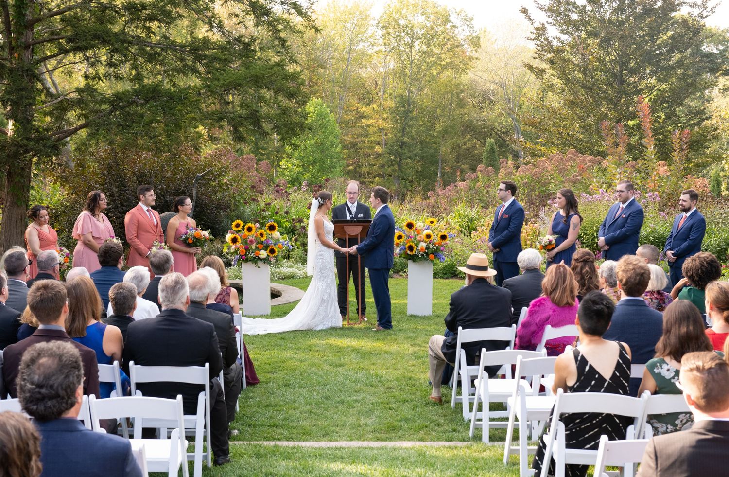 Wedding ceremony guests seated in white chairs outdoors during golden hour sunset in a garden setting.
