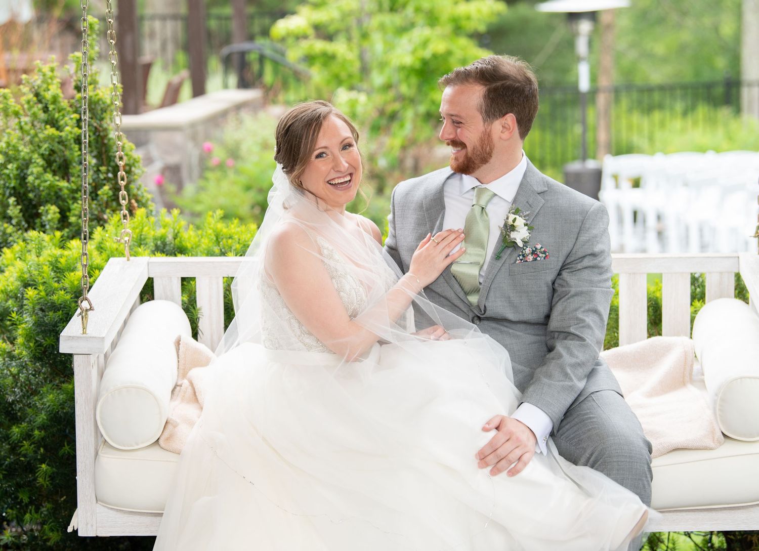 A couple in wedding attire shares joyful moments on a white porch swing during their outdoor celebration.
