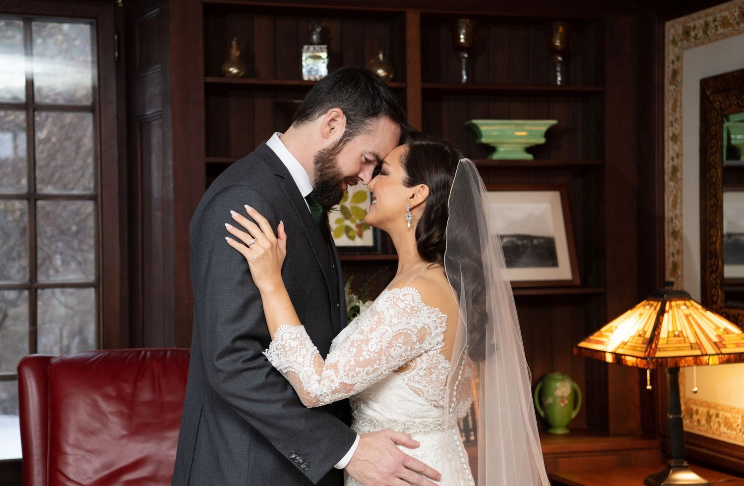 A romantic wedding couple shares an intimate kiss in a dimly lit room with windows and warm lighting.