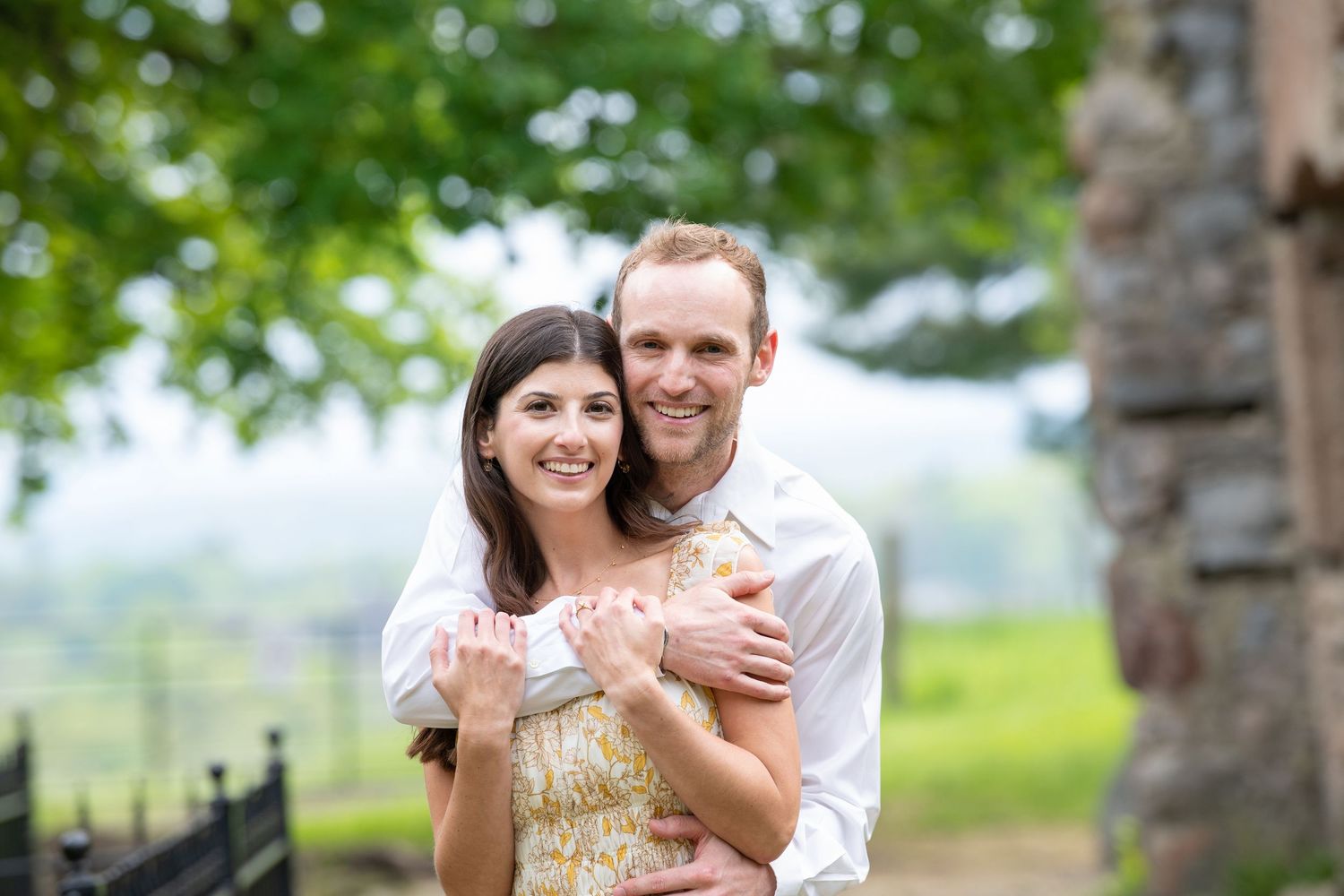 A happy couple embraces outdoors during their wedding photoshoot with a blurred green landscape in the background.