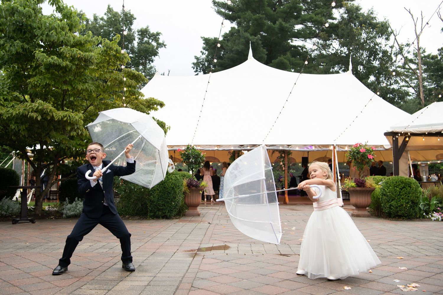 Two people in wedding attire playfully pose with clear umbrellas in front of a white tent at an outdoor venue.