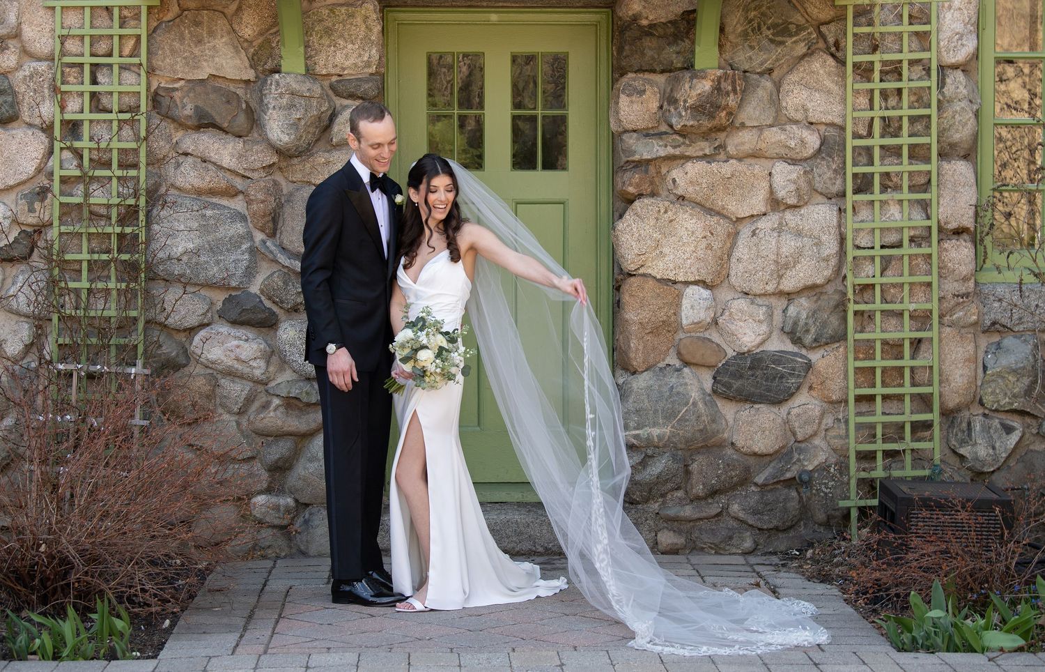Bride and groom pose before stone wall with green lattice door, bride holding bouquet with flowing veil.