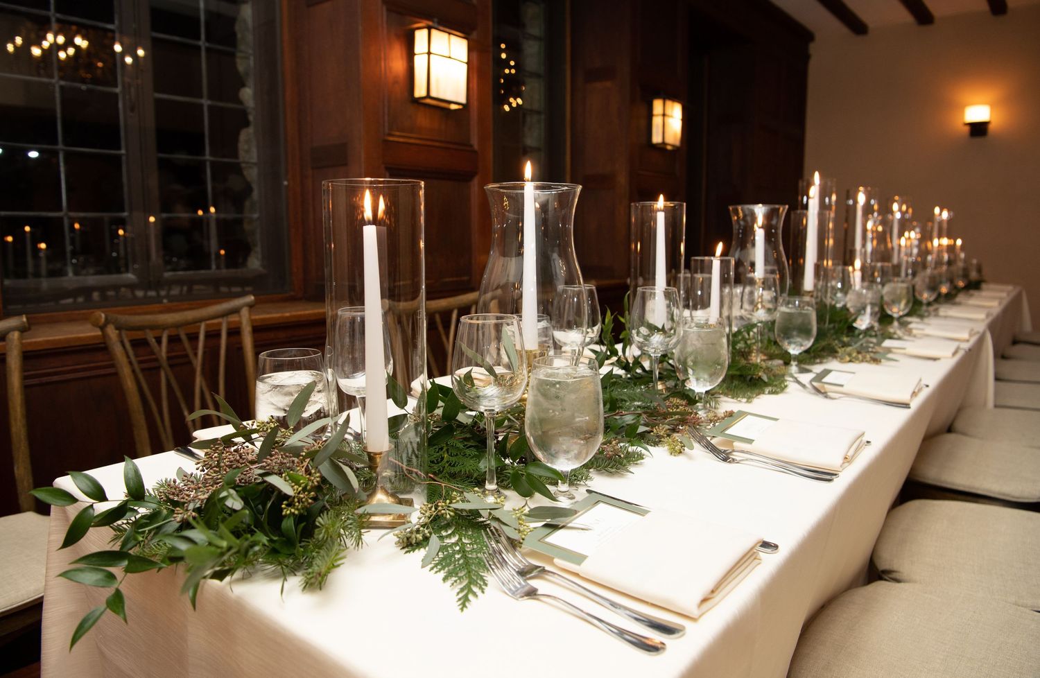 An elegant wedding reception table with white linens, crystal glassware and fresh greenery garland centerpieces.