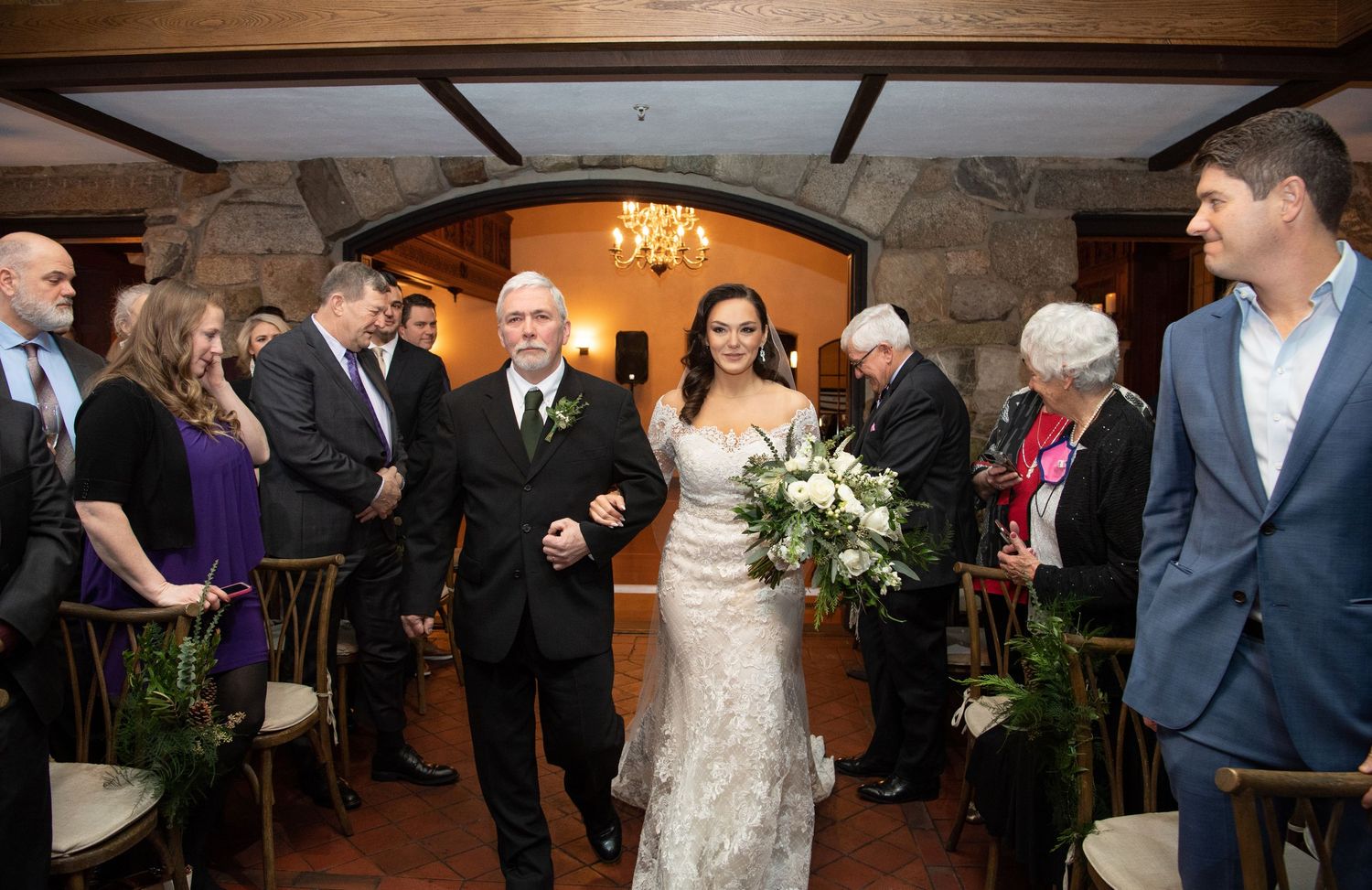 A wedding ceremony takes place in an elegant indoor venue with exposed beams and a chandelier overhead.