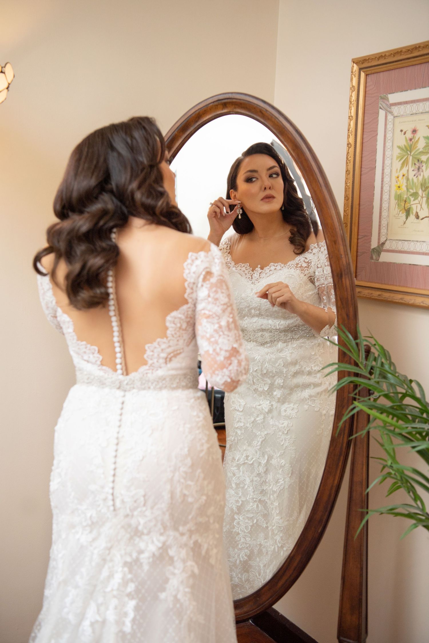 A bride in an elegant lace wedding gown with sheer sleeves looks at her reflection in an oval wooden-framed mirror.