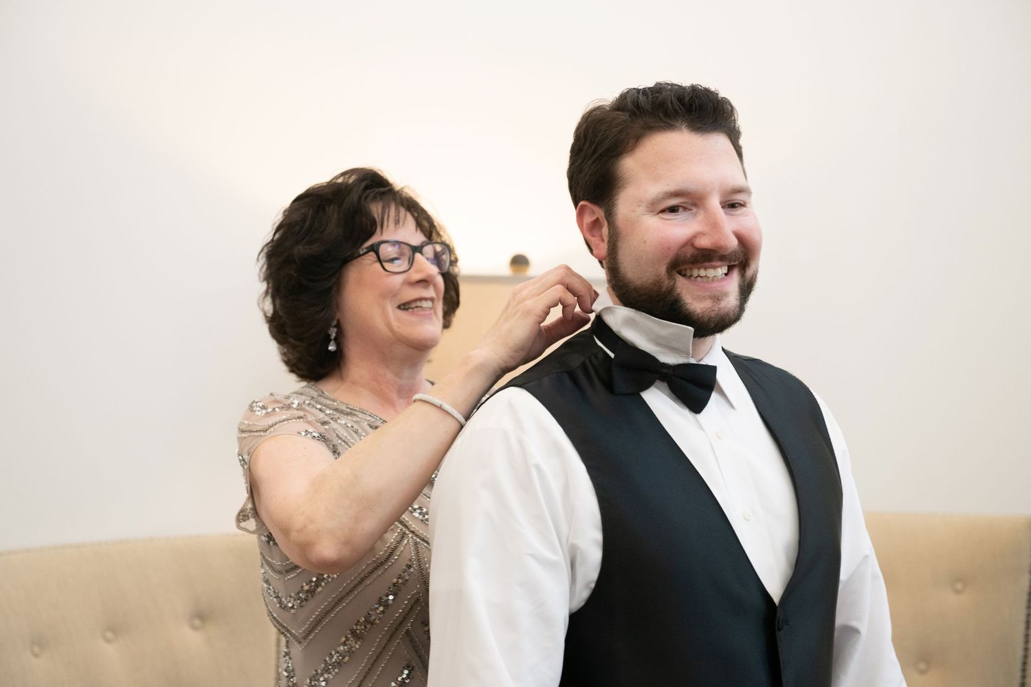 Two people share a joyful moment on a beige couch as one adjusts the other's bowtie while both laugh.