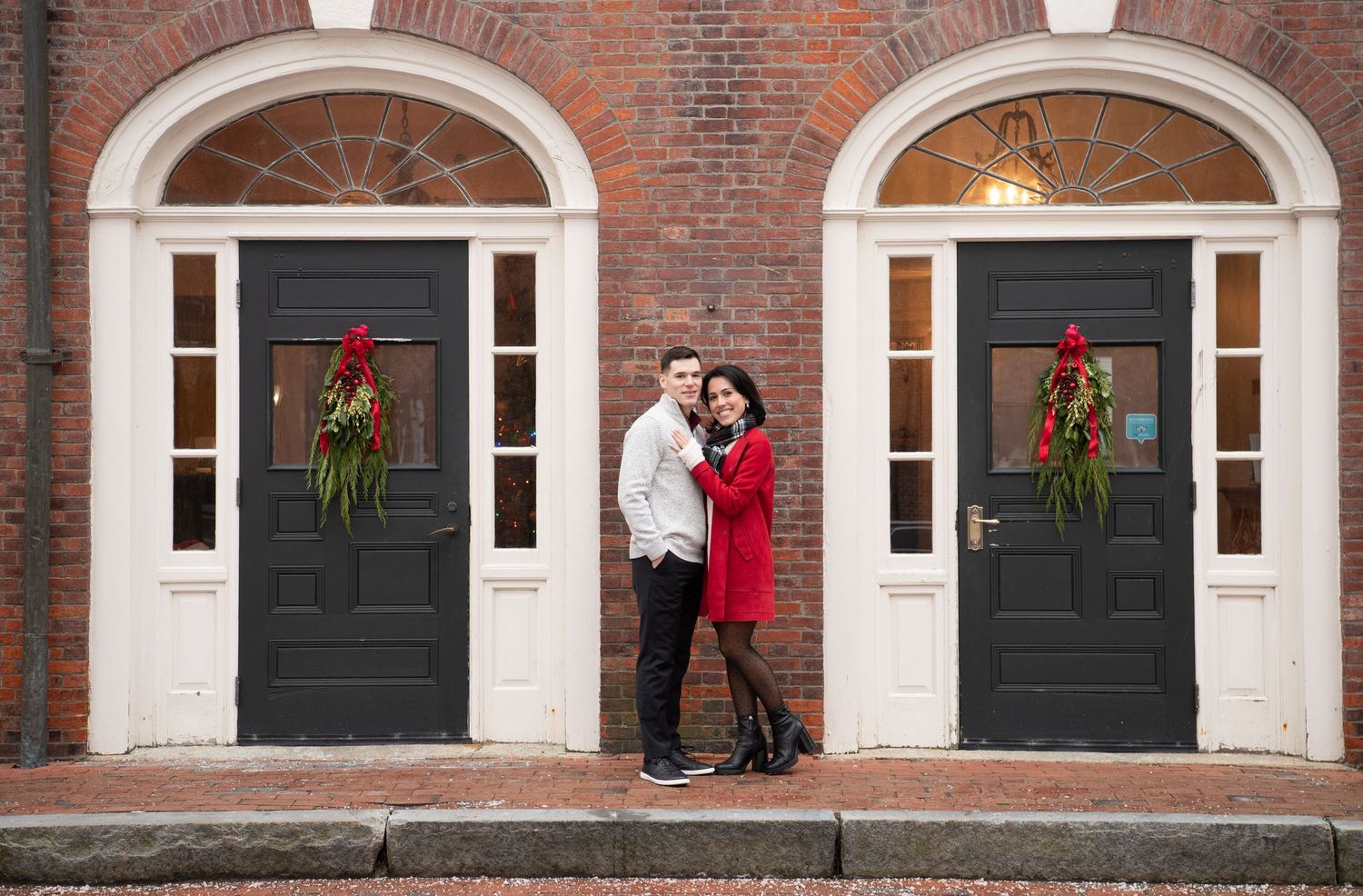 A couple embraces in front of a classic brick building with arched doorways decorated with holiday wreaths.