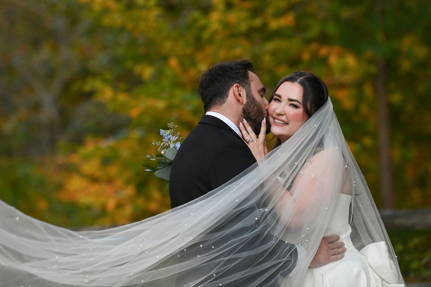 A romantic wedding couple embraces in a wooded setting with vibrant autumn foliage in the background.