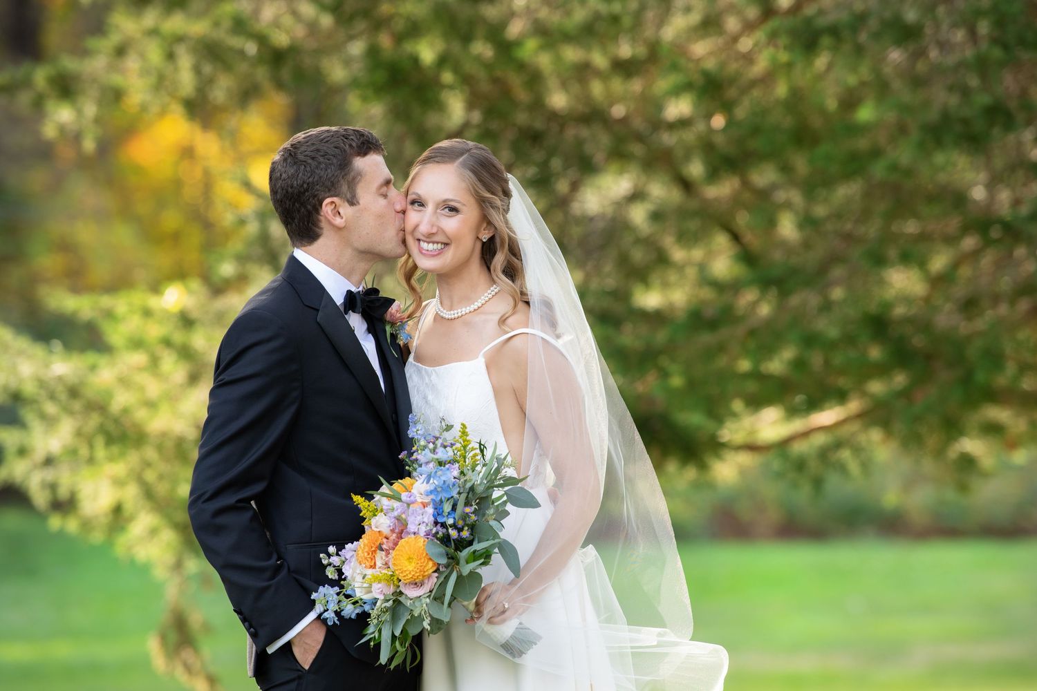 A bride in a white gown holds a colorful bouquet while standing with groom in black suit outdoors in a garden setting.