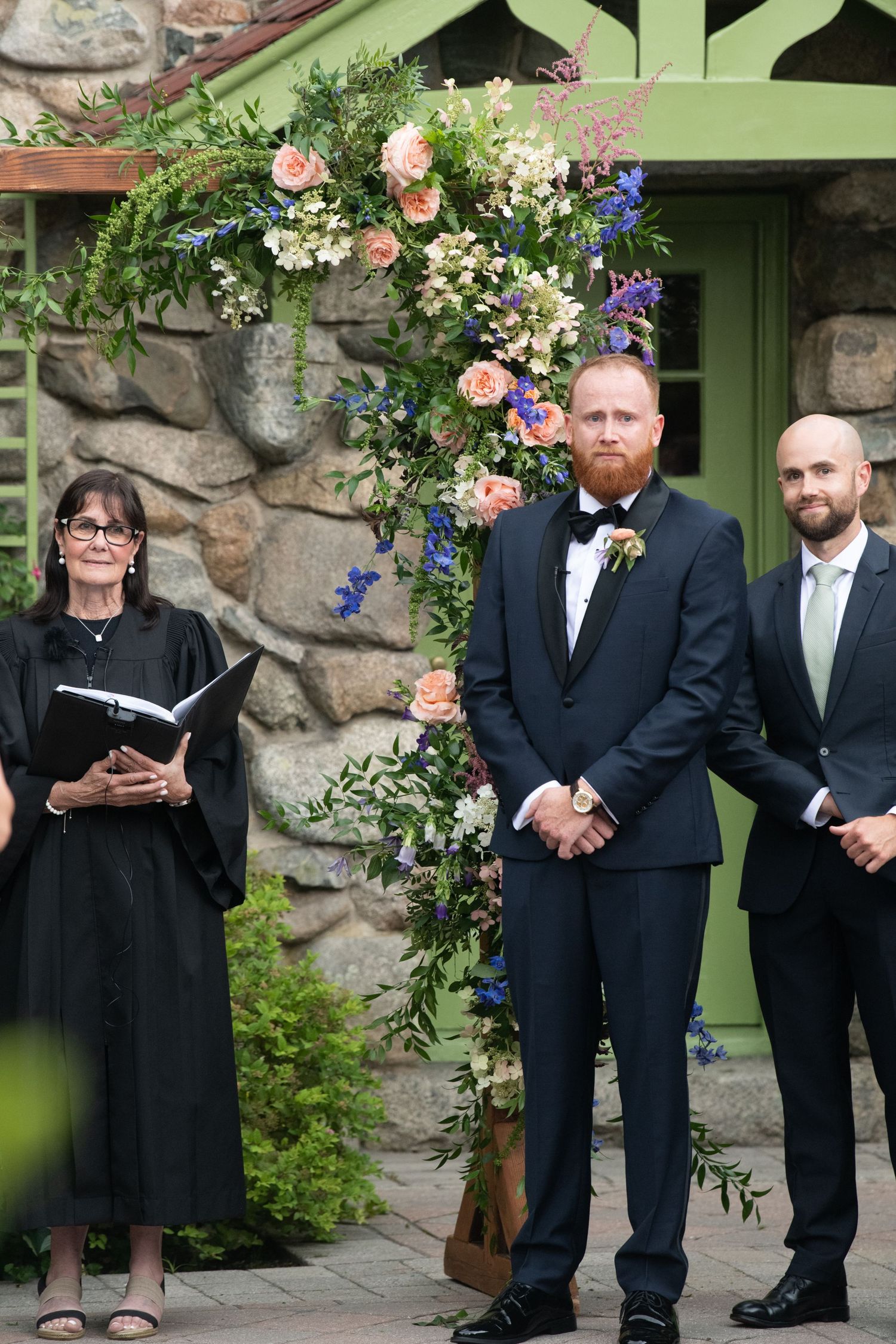 Outdoor wedding ceremony with officiant and wedding party under floral arch against stone wall.