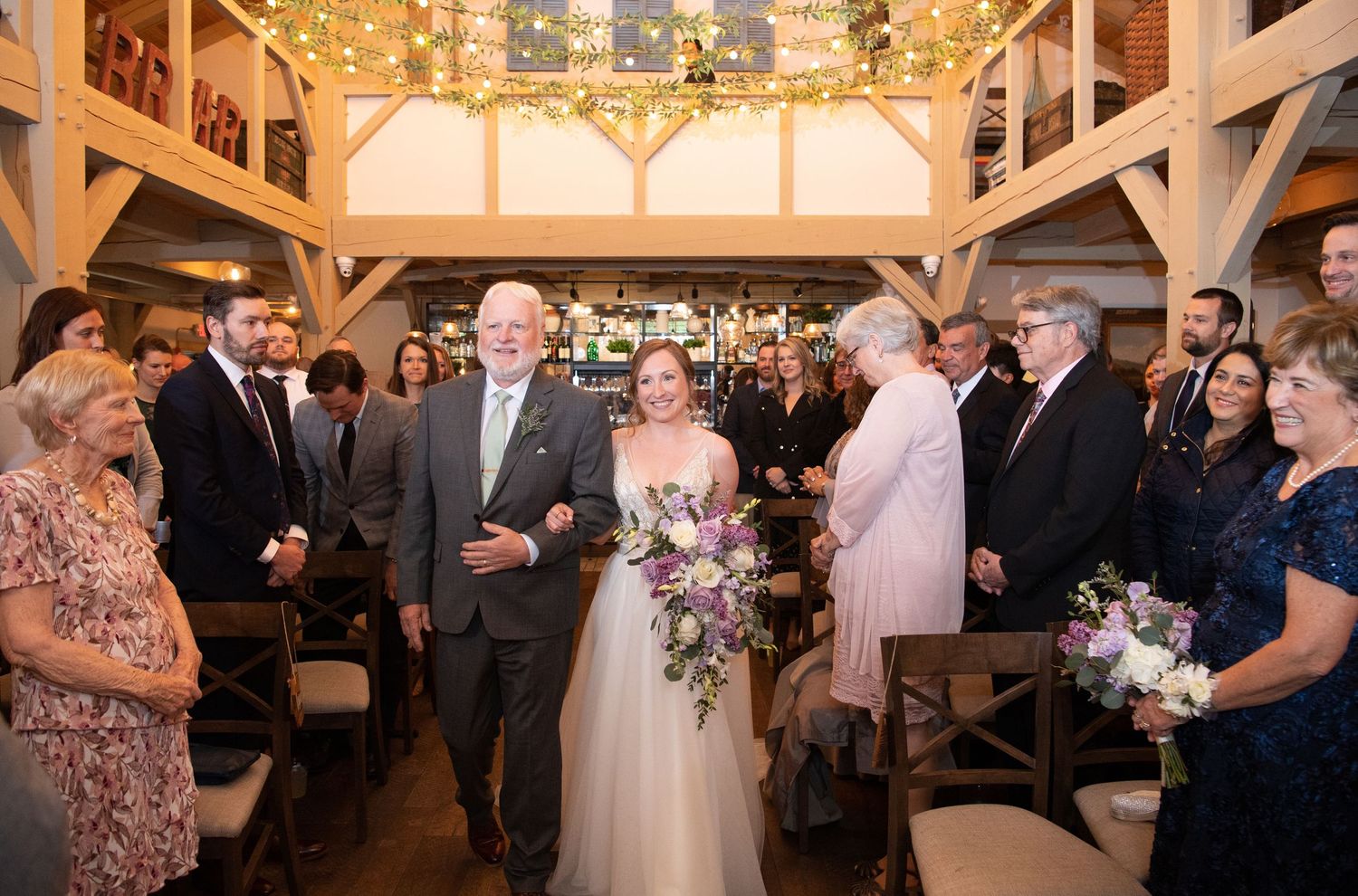 Wedding reception guests mingle and celebrate in an elegantly decorated rustic barn venue with wooden beams and warm lighting.