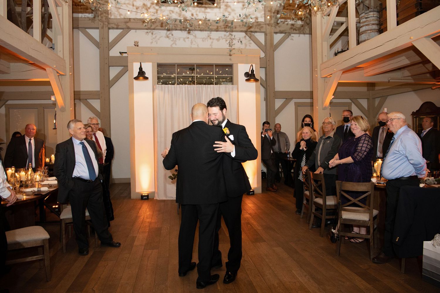Wedding guests mingle and socialize in an elegant white-walled reception venue with wooden beam accents.