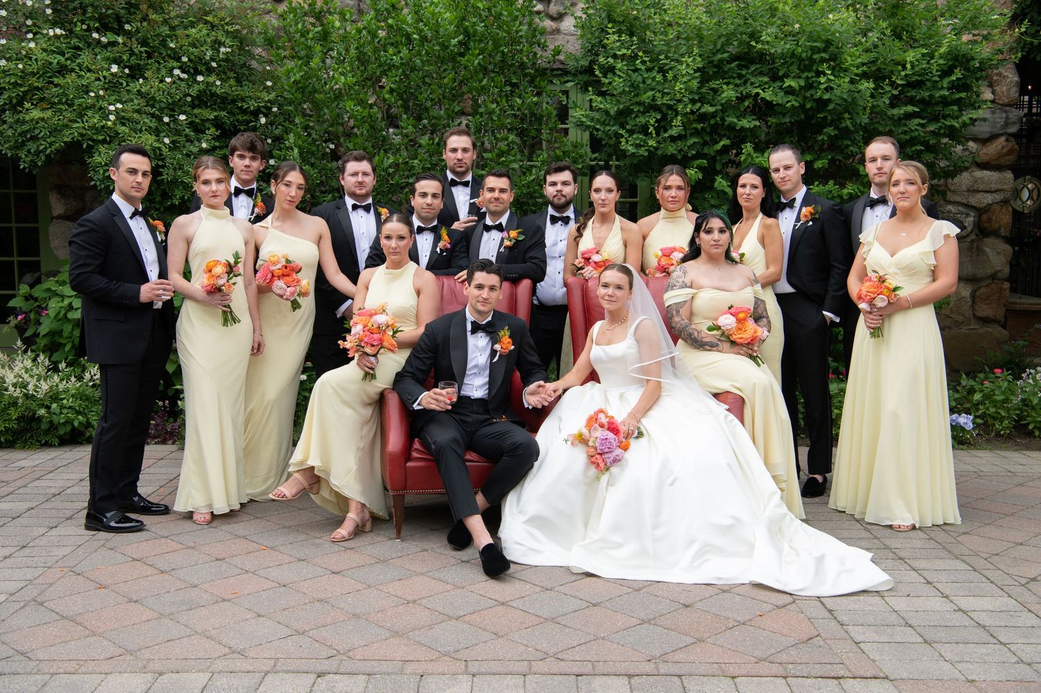 Wedding party poses outdoors, bride and groom seated, bridesmaids in cream gowns holding coral bouquets, groomsmen in black tuxedos.