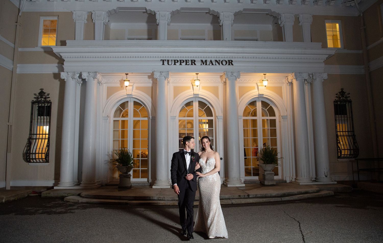Bride and groom pose romantically at night in front of the elegantly lit Tupper Manor wedding venue.