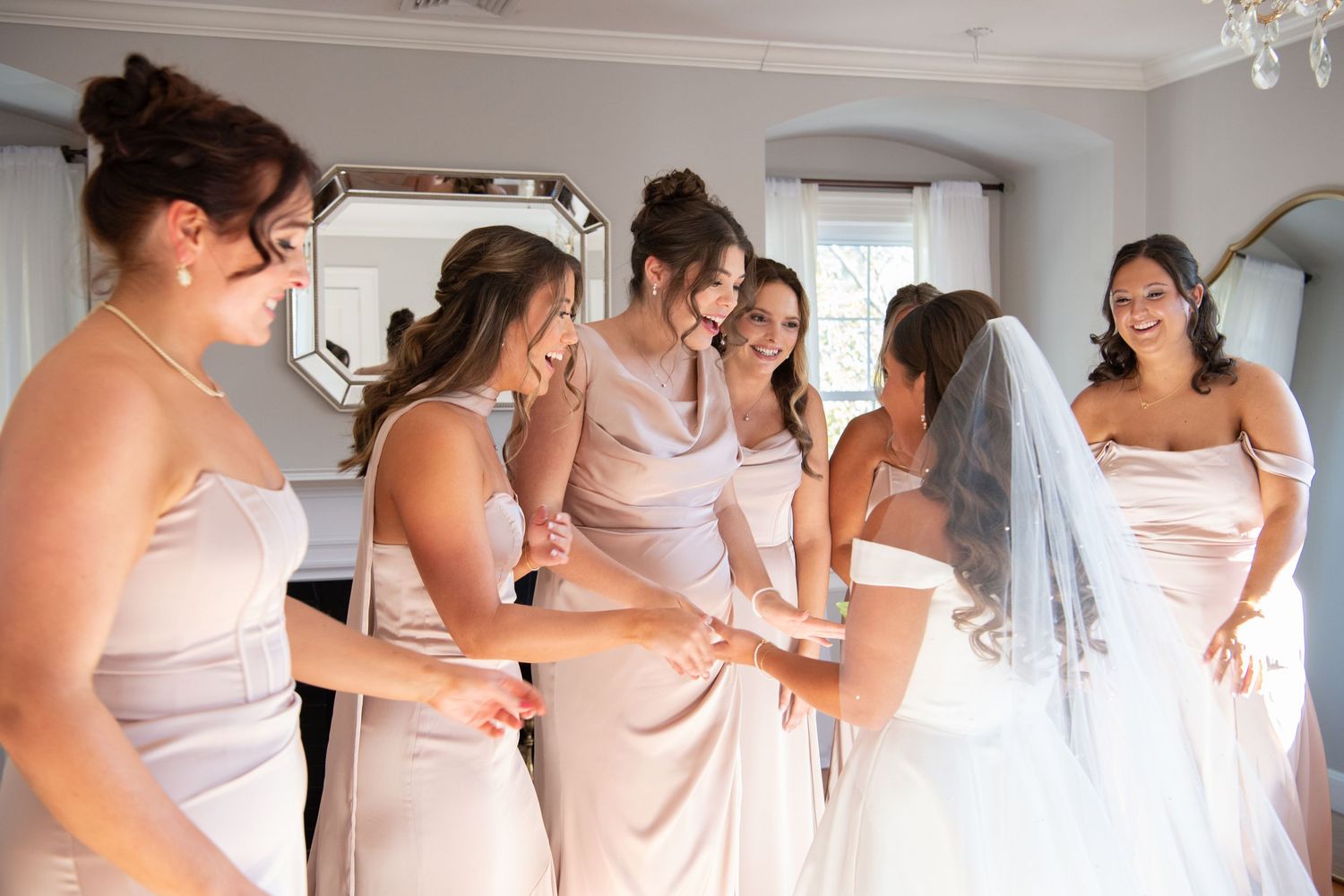 Bridesmaids in blush pink dresses help the bride get ready in a bright white room.